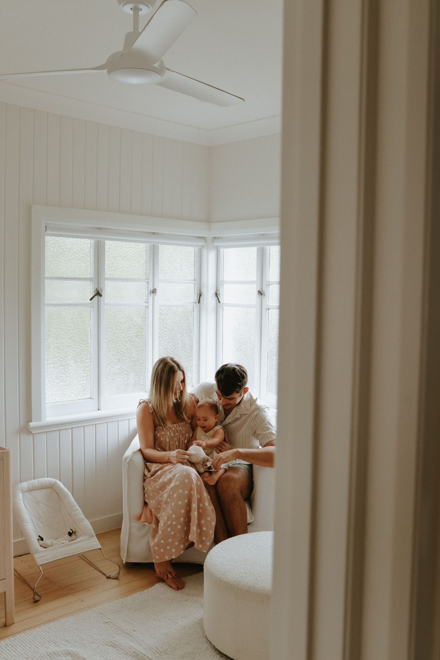 Intimate family moment in a bright sunlit window nook with white walls and ceiling fan.