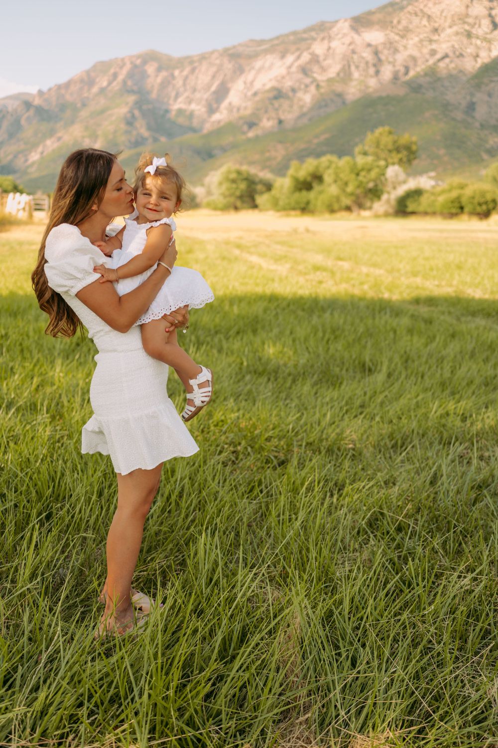 A mother lifts her young daughter in a grassy field with mountains in the background.
