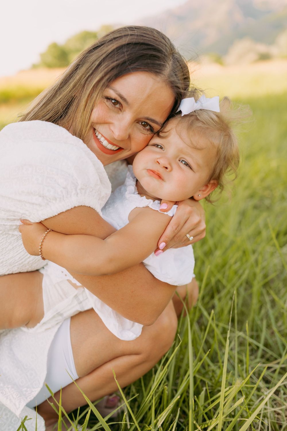 A tender embrace between parent and child wearing white dresses in a sunny meadow with mountains in the background.