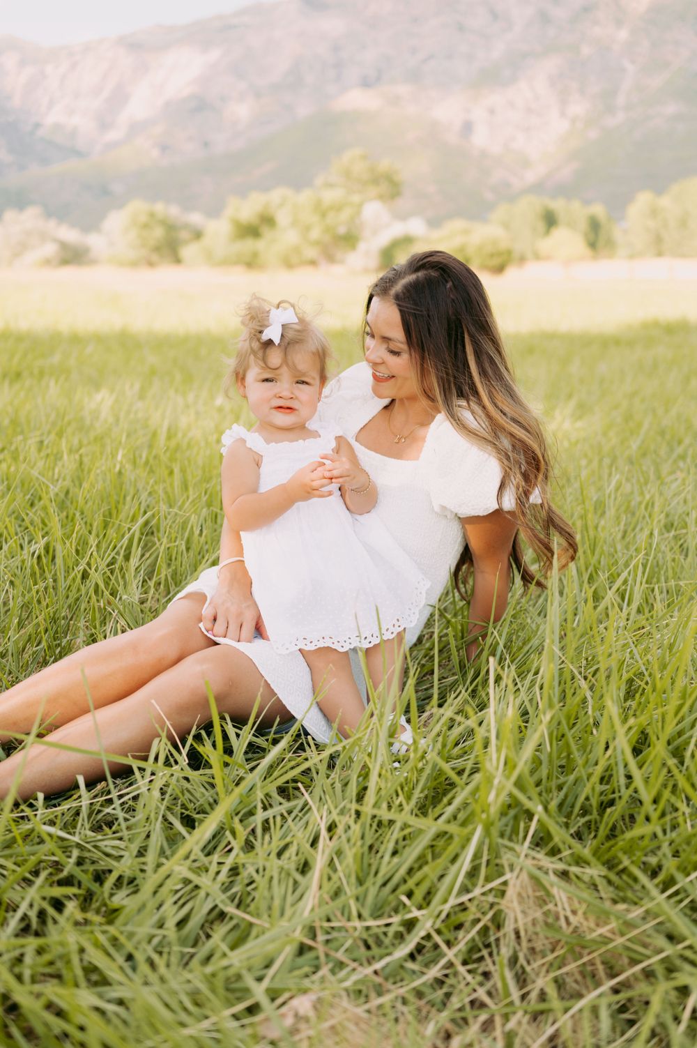 Sweet bonding moment between parent and toddler sitting in tall grass with mountain scenery.