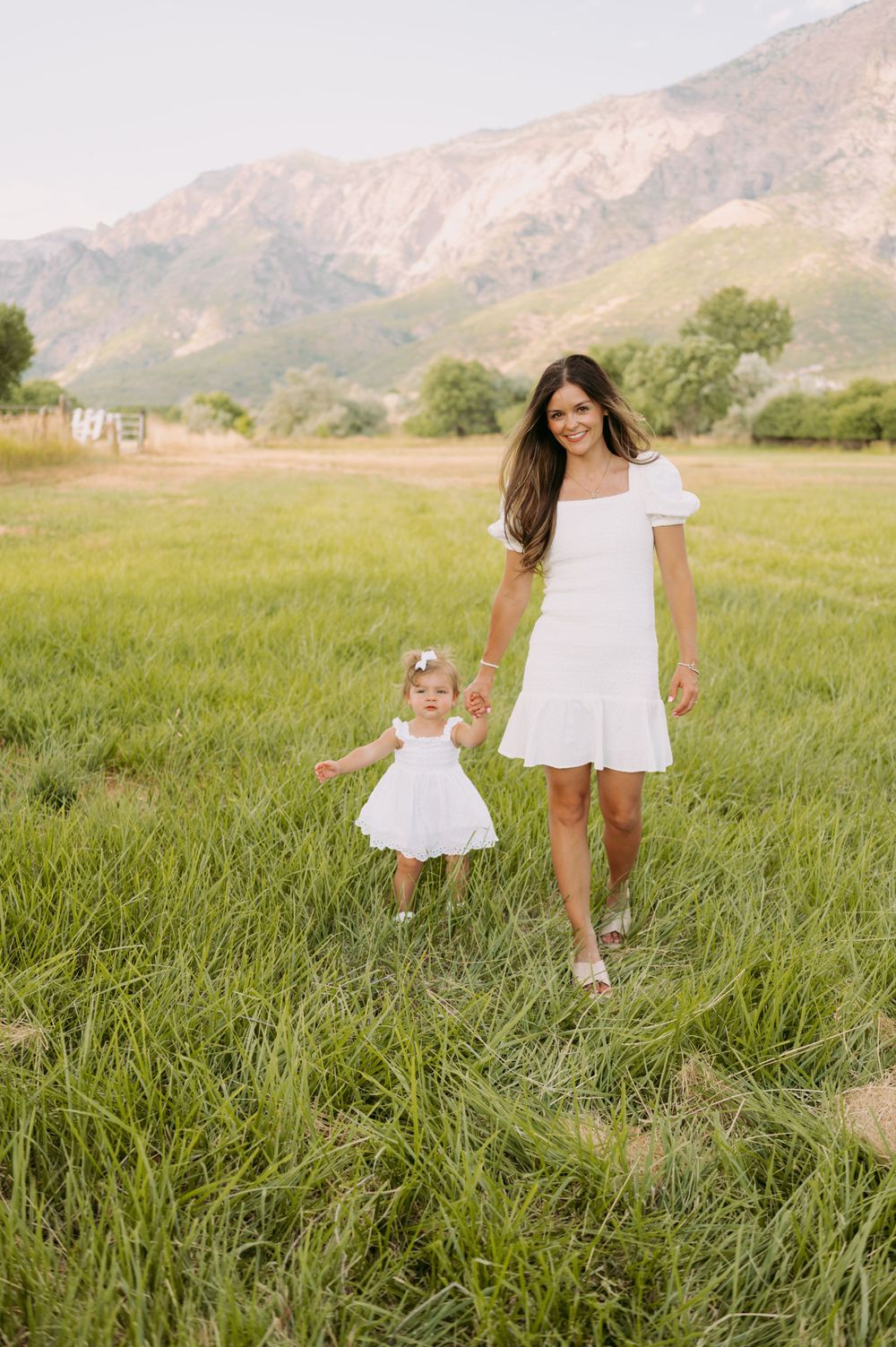 Parent and child in matching white dresses walking hand in hand through a mountain meadow.