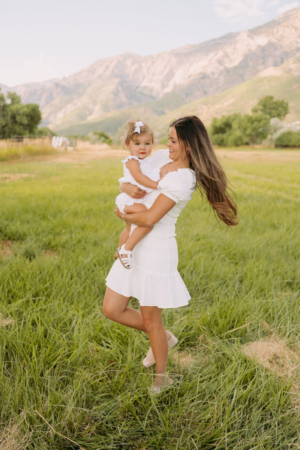 Parent holding child in coordinated white outfits pose in a grassy field with mountains behind them.