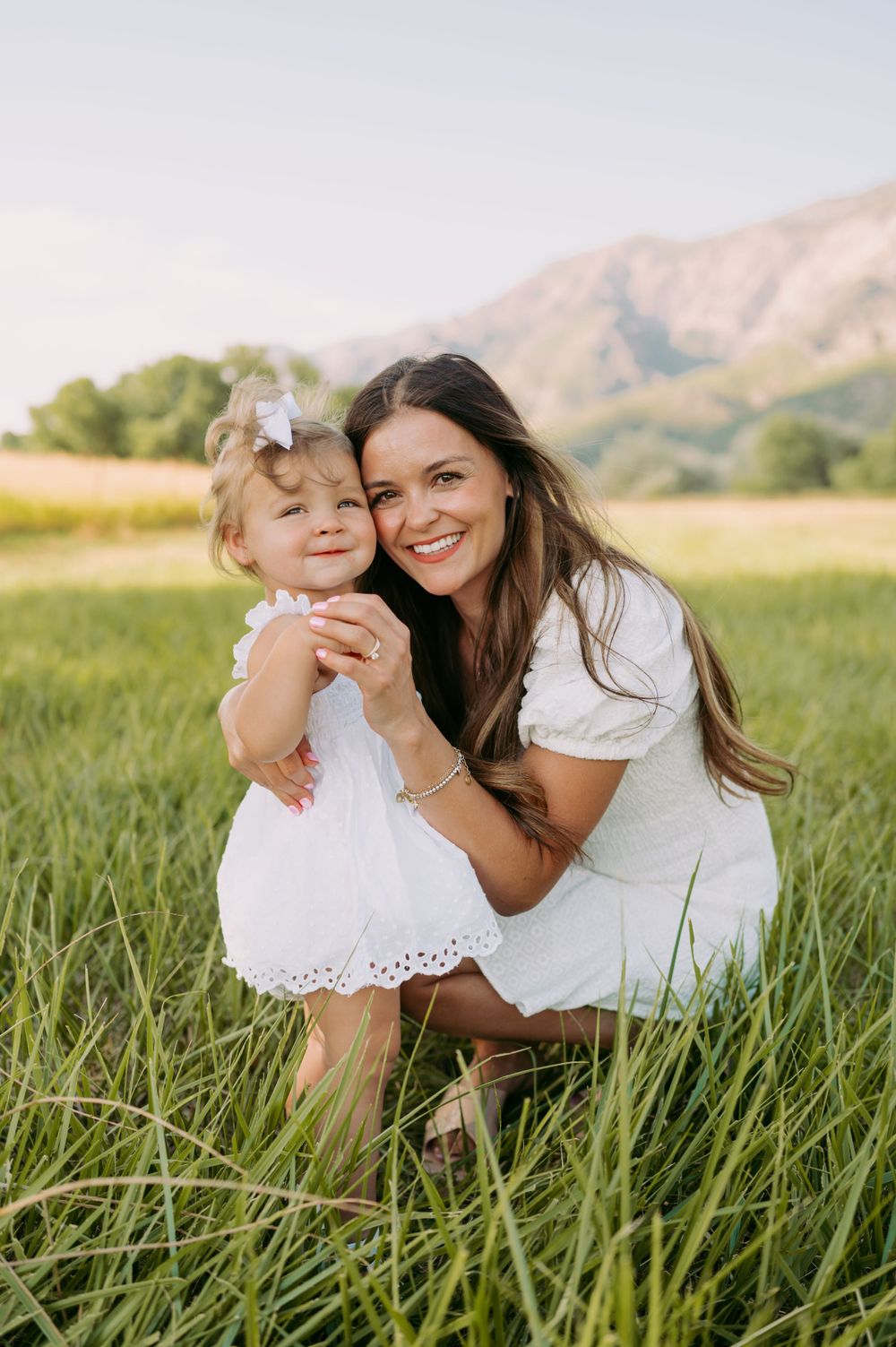 A tender moment between mother and daughter in white dresses against a mountain backdrop.