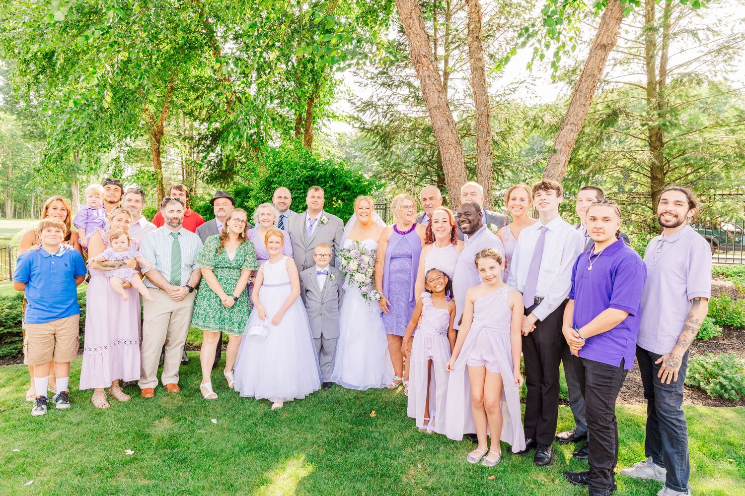 Large wedding party poses outdoors under green trees on a sunny summer day.