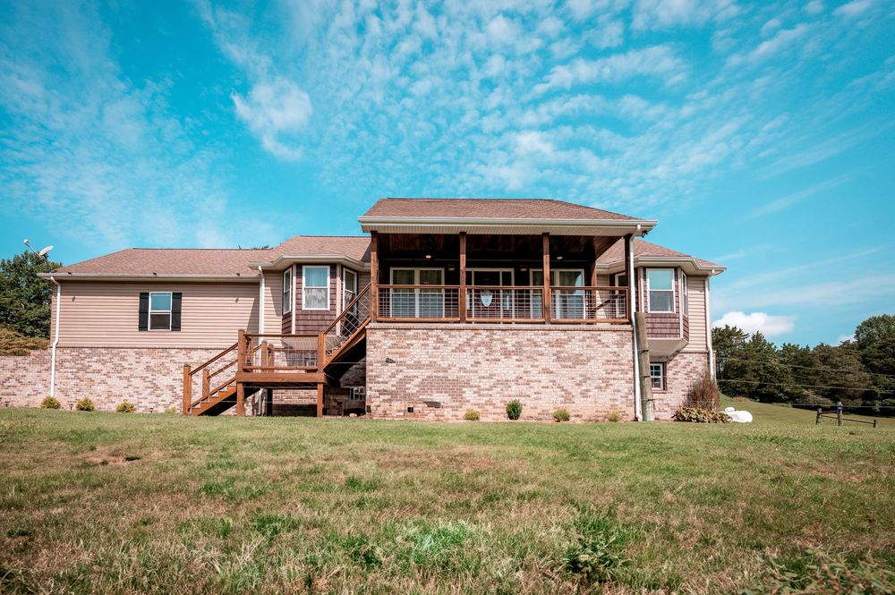 Panoramic view of a ranch-style brick house with a wraparound porch set against a bright blue sky on rolling hills.