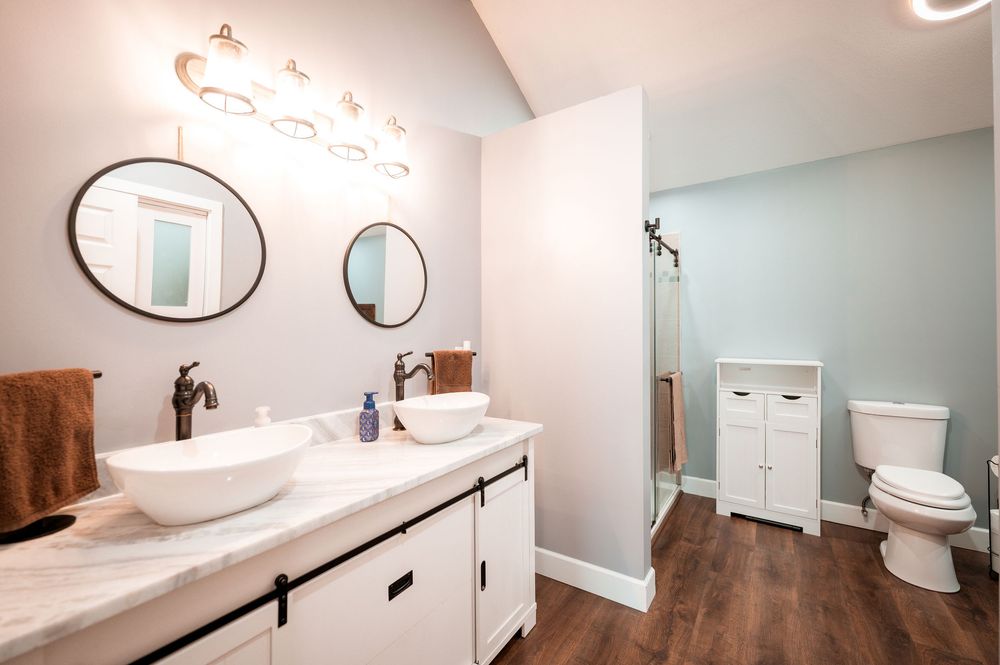 Modern bathroom renovation features white vanity, oval mirrors, hardwood floors, and sleek fixtures in a clean design.