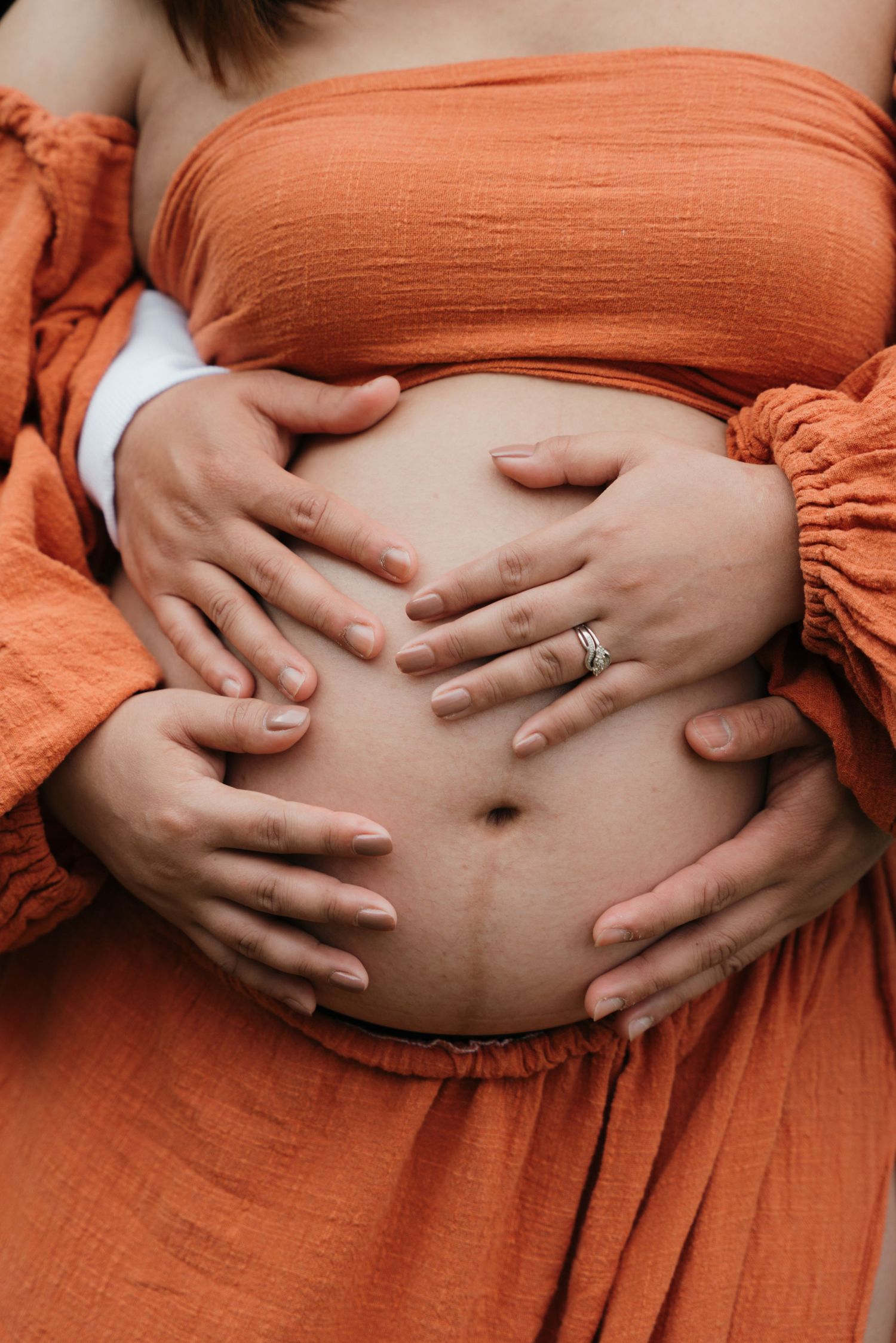 Close-up of pregnant belly with hands forming heart shape, wearing orange dress.