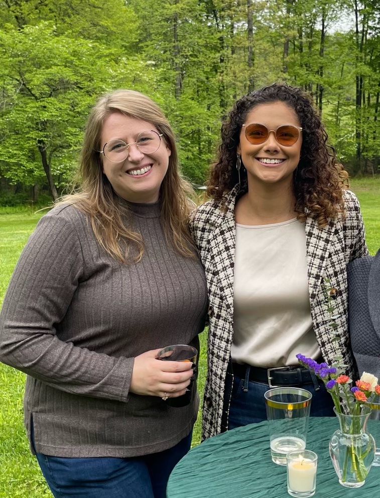 Two friends enjoy drinks and conversation at an outdoor gathering with greenery in background.