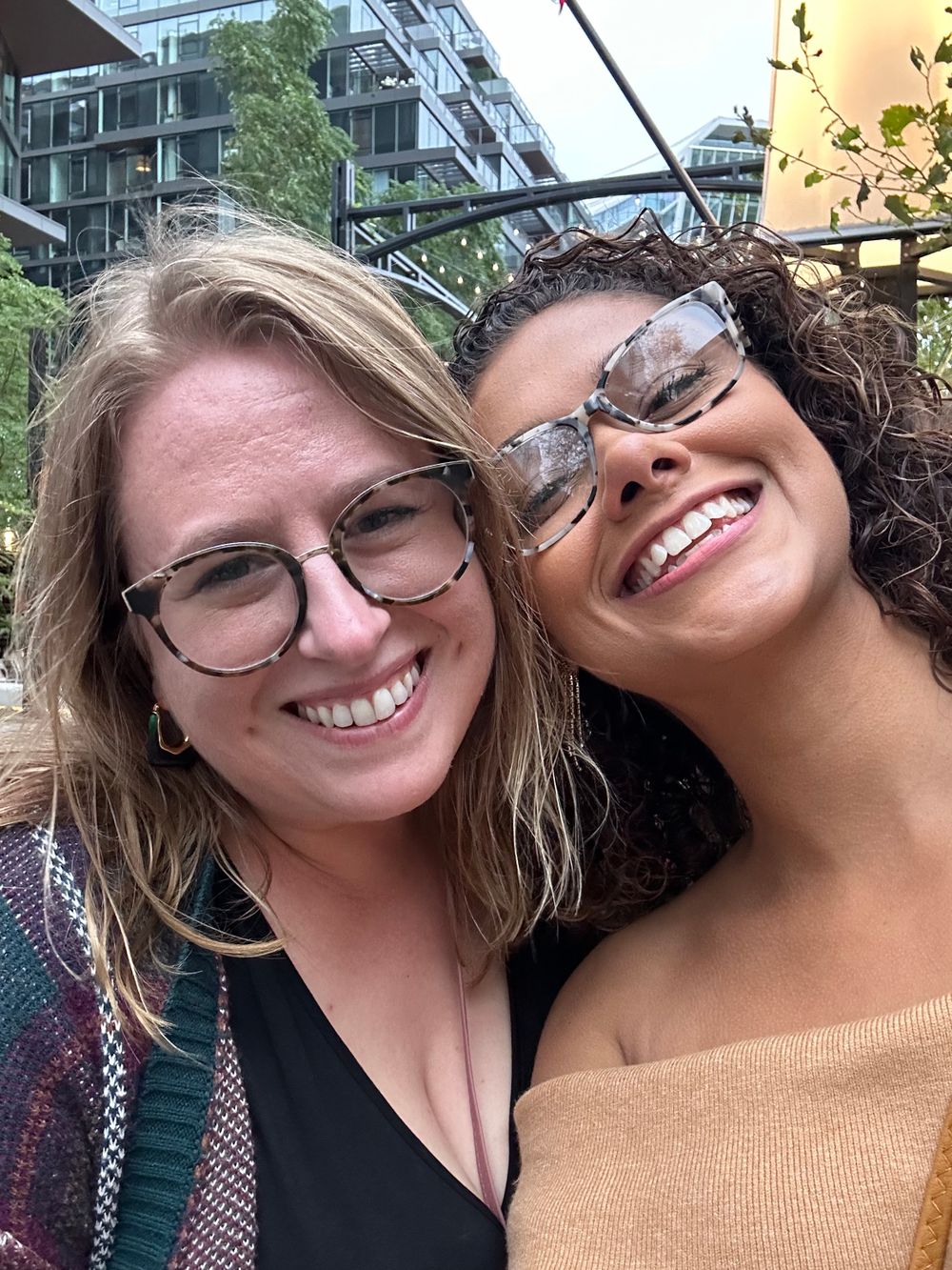 Two friends take a cheerful selfie together in front of modern apartment buildings.