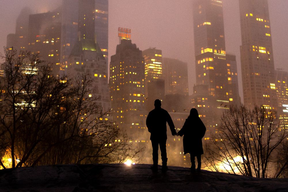 Silhouettes of people stand against a foggy city skyline at night with glowing buildings in the background.