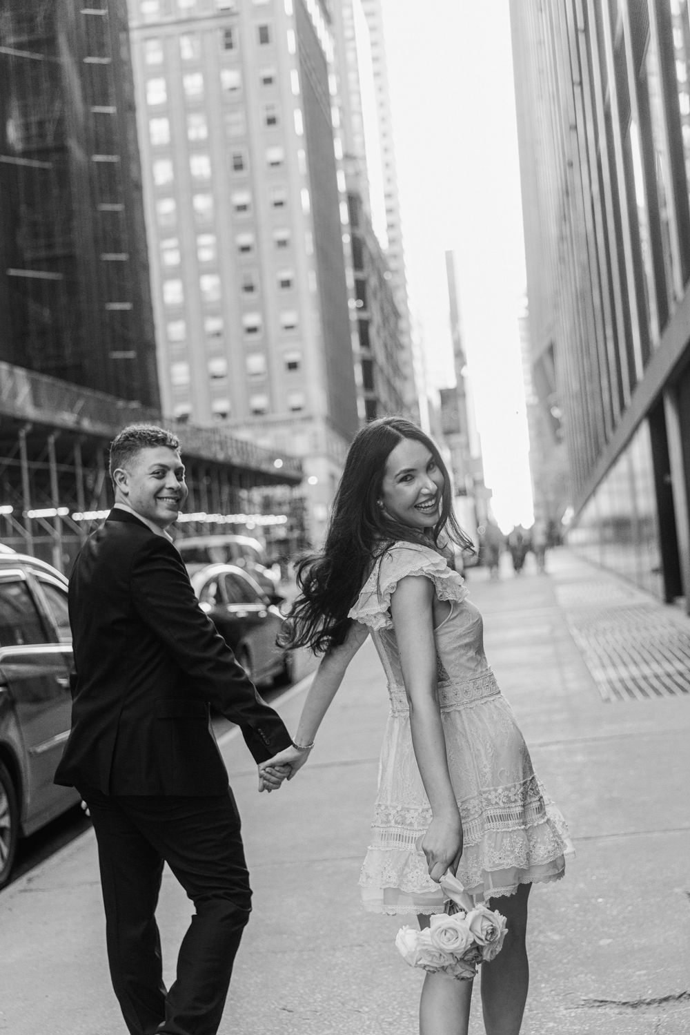 Black and white photo of a smiling couple holding hands on a city sidewalk, woman in white dress holding bouquet.
