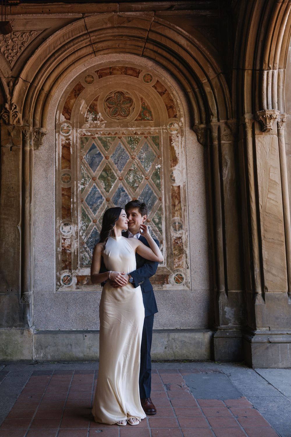 Couple embracing under ornate stone arch at Bethesda Terrace, Central Park, woman in elegant ivory gown.