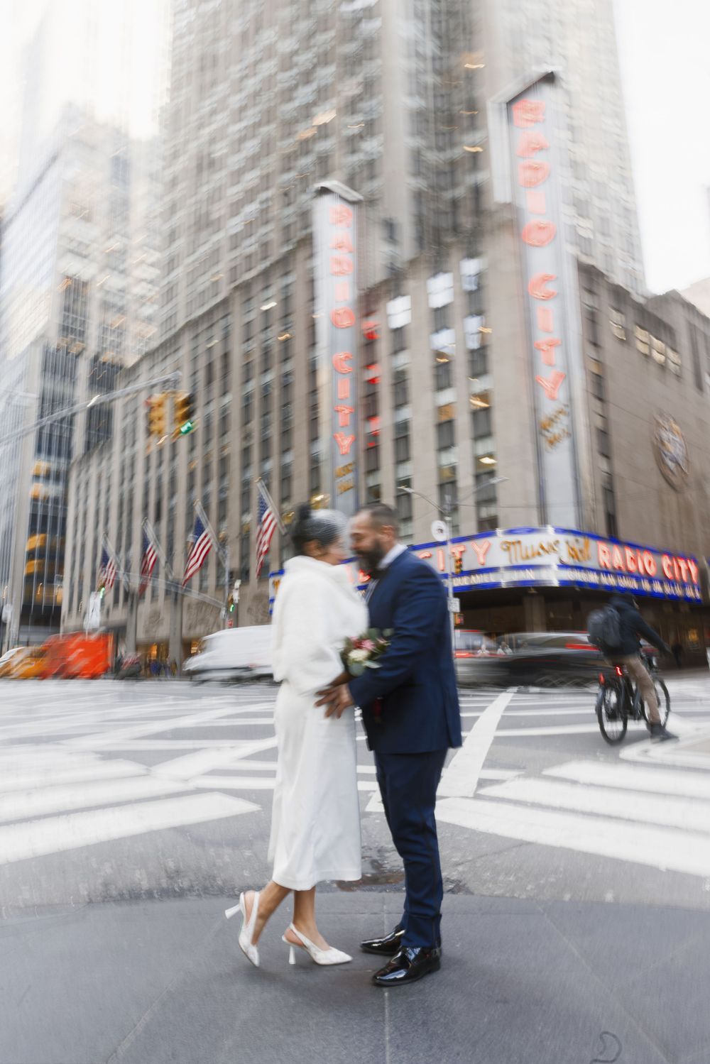 A romantic couple in wedding attire dances and spins on a city street with tall buildings in the background.