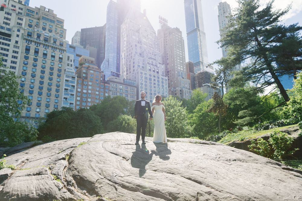 A bride and groom stand on a rock in Central Park with NYC skyline skyscrapers and lush trees in the background.