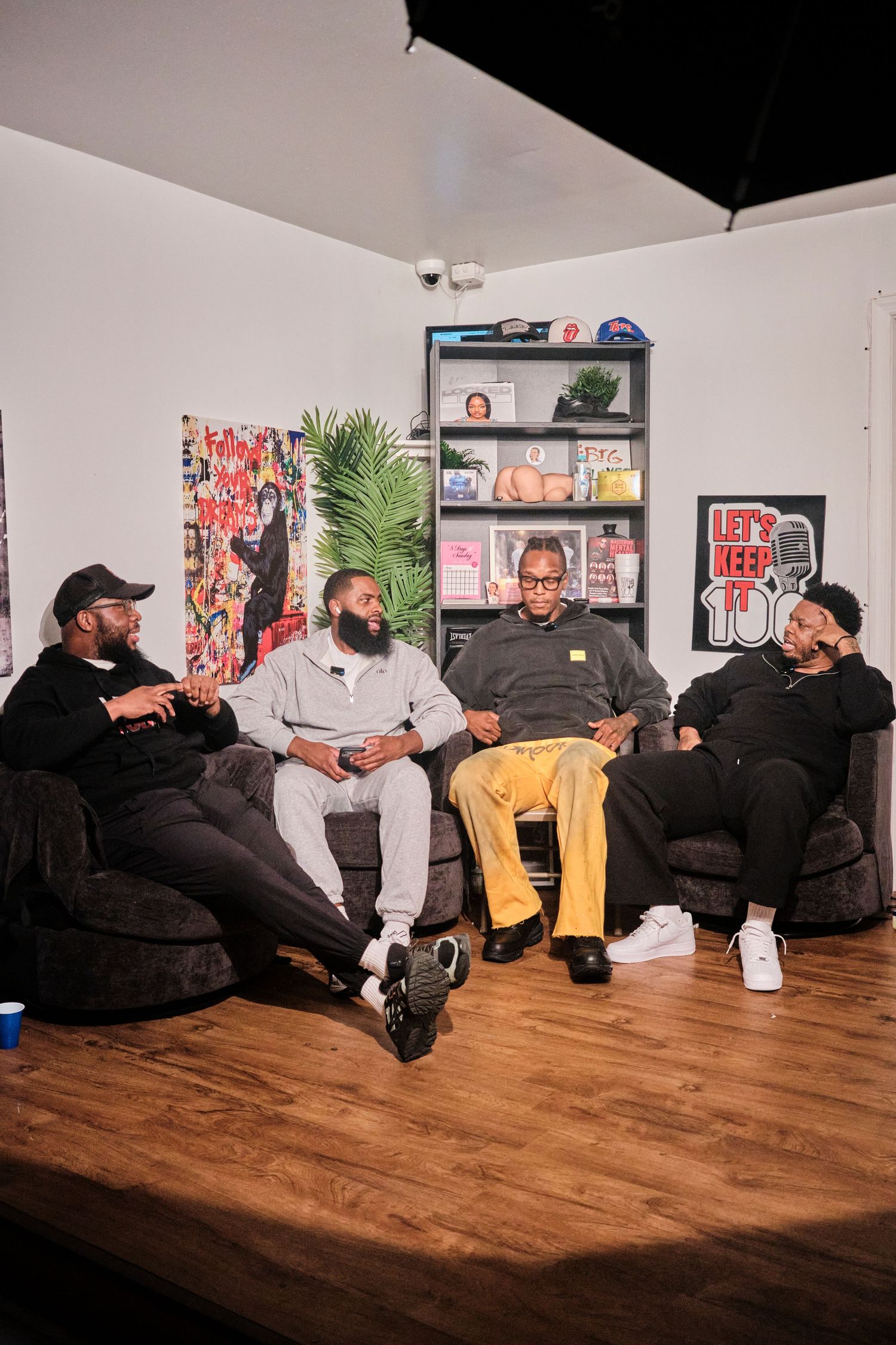 Four men sitting on couches in a podcast studio with artwork and a bookshelf in the background.