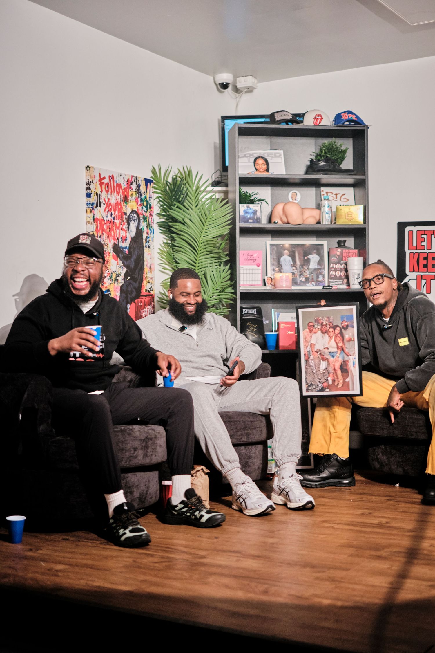 Three men laughing and sitting on a dark couch in a casual podcast studio setting with shelves and artwork behind them.