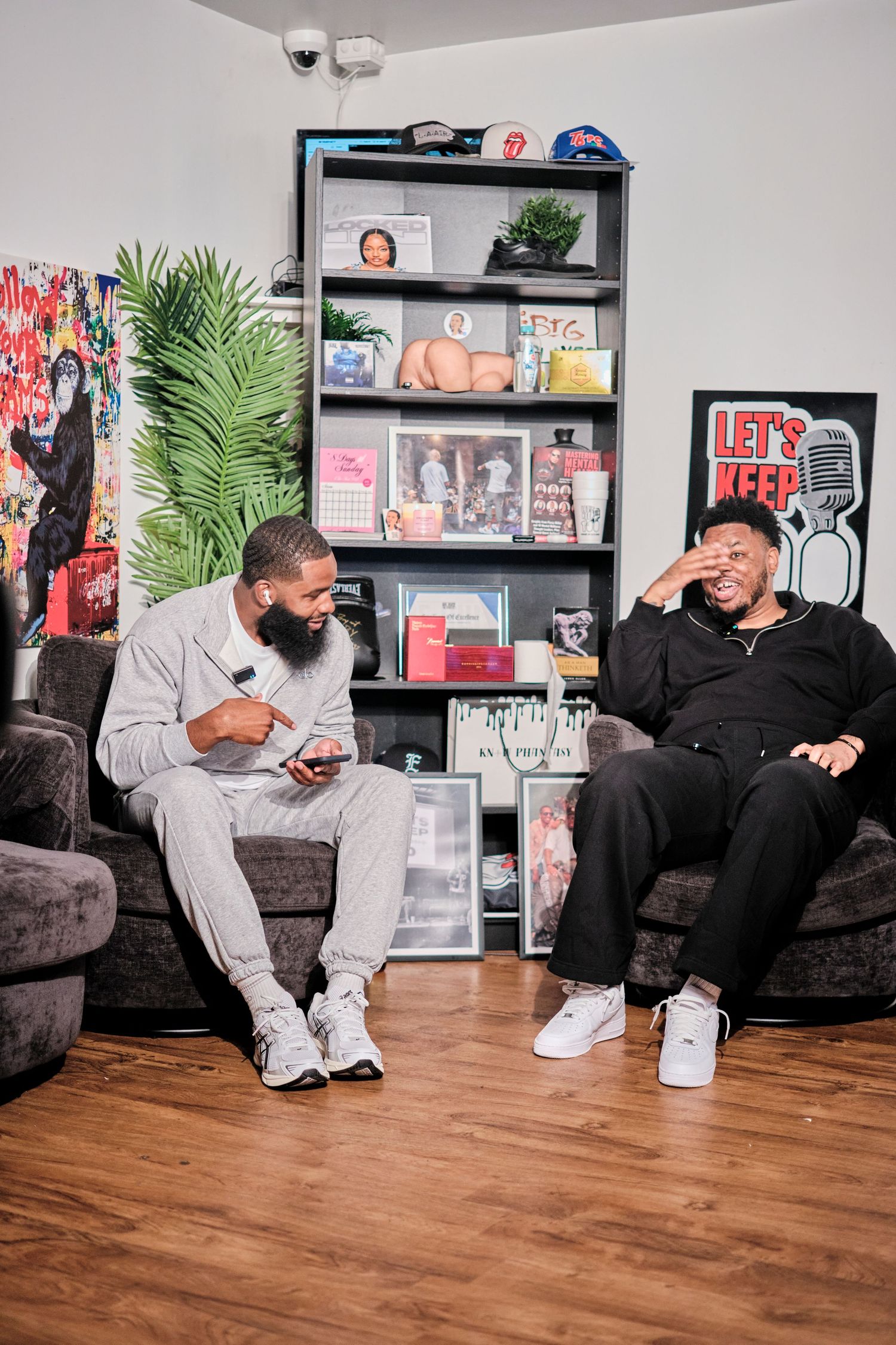 Two men laughing and conversing on dark sofas in a stylish room with shelves and a Let's Keep It Real podcast sign.