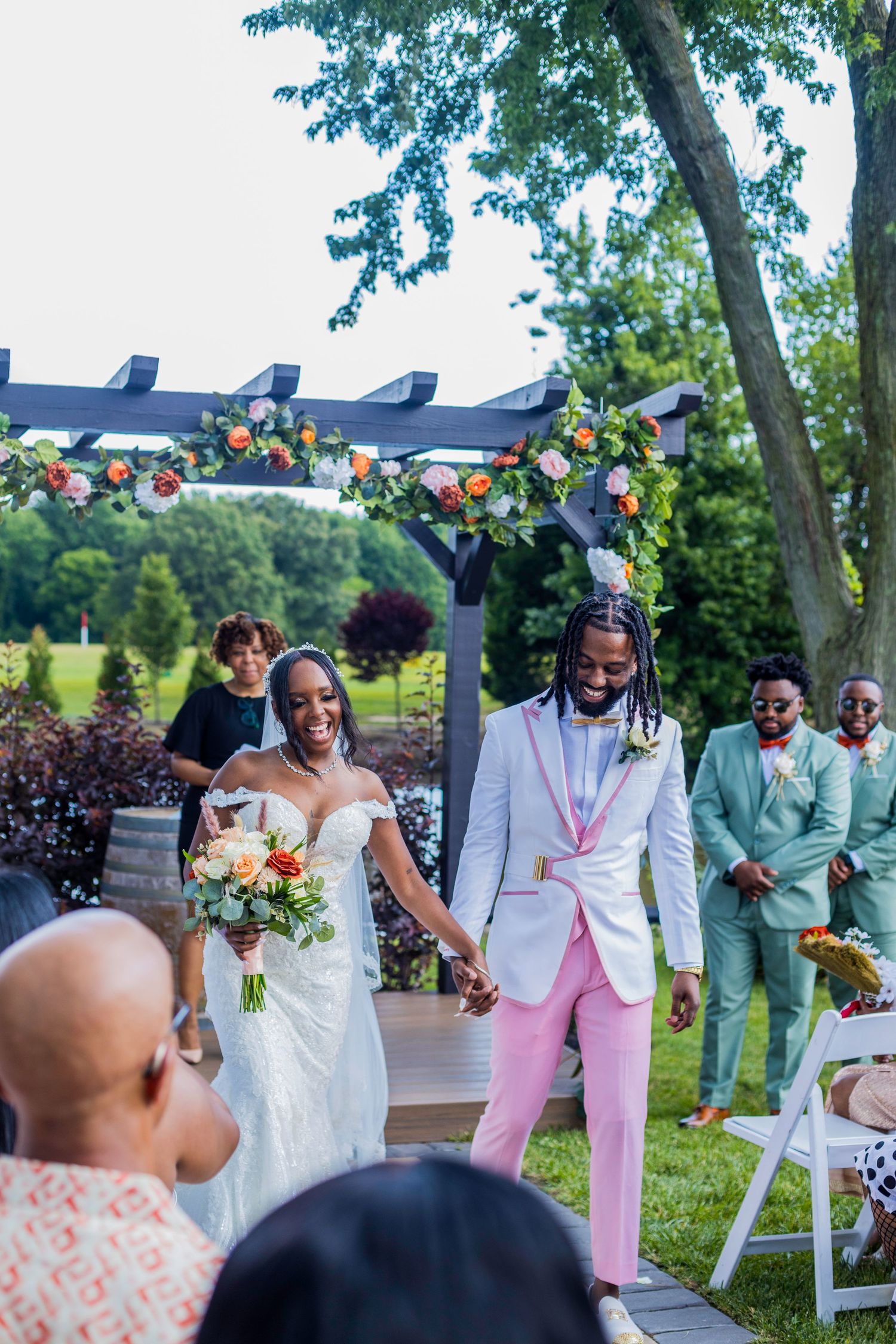 Wedding party celebrates joyfully outdoors with bridesmaids in pink dresses and groomsmen in white suits.
