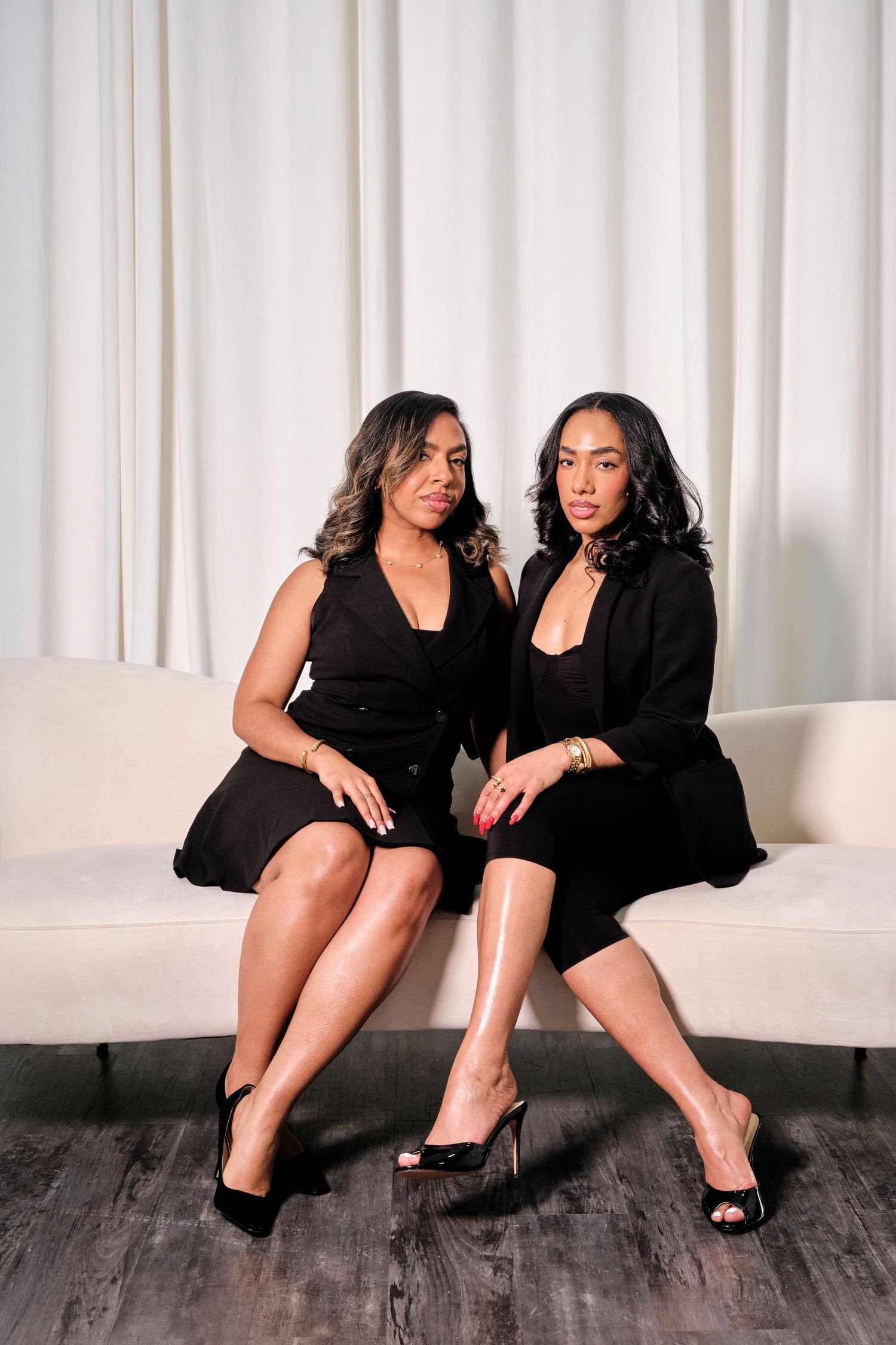 Two women in black dresses sitting together on a white sofa against a white curtain backdrop.