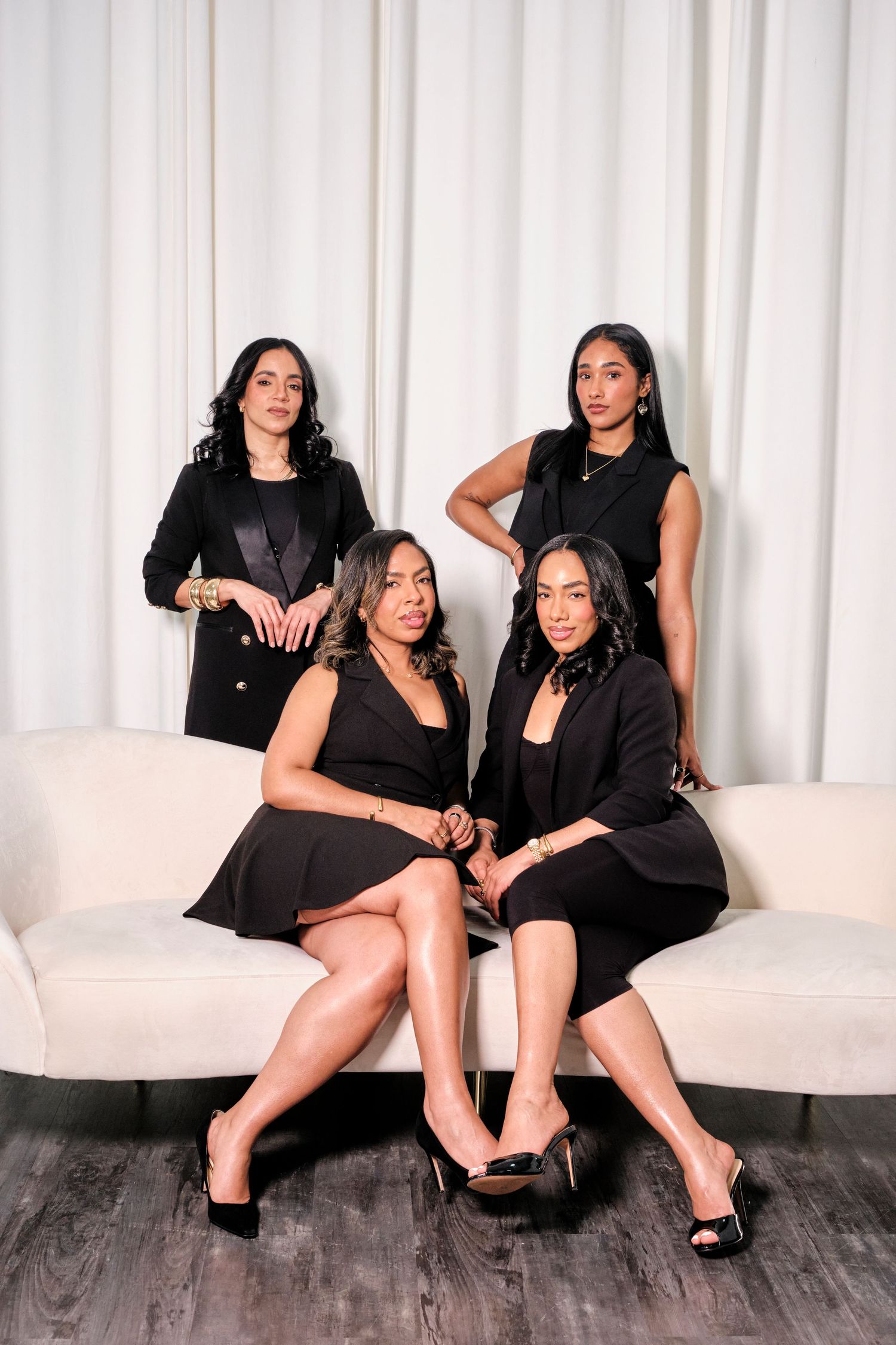 Four women in matching black outfits pose elegantly on a white sofa against a white curtain backdrop.