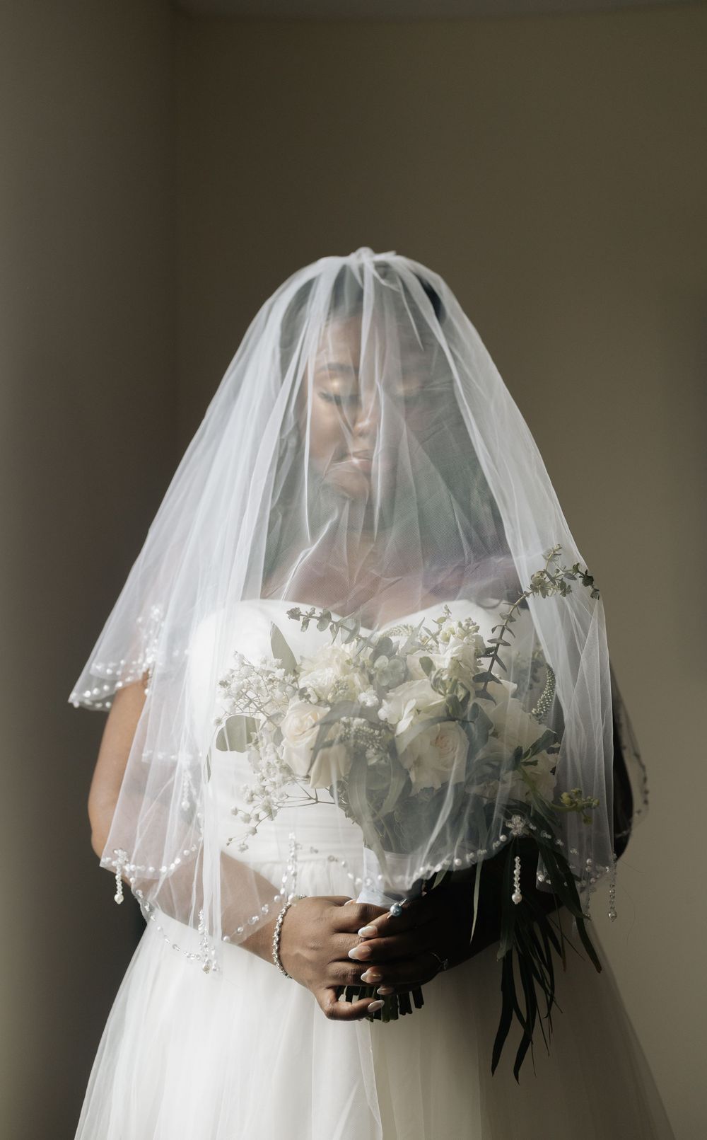 Elegant bride wearing traditional white gown and veil holds bouquet in moody portrait.