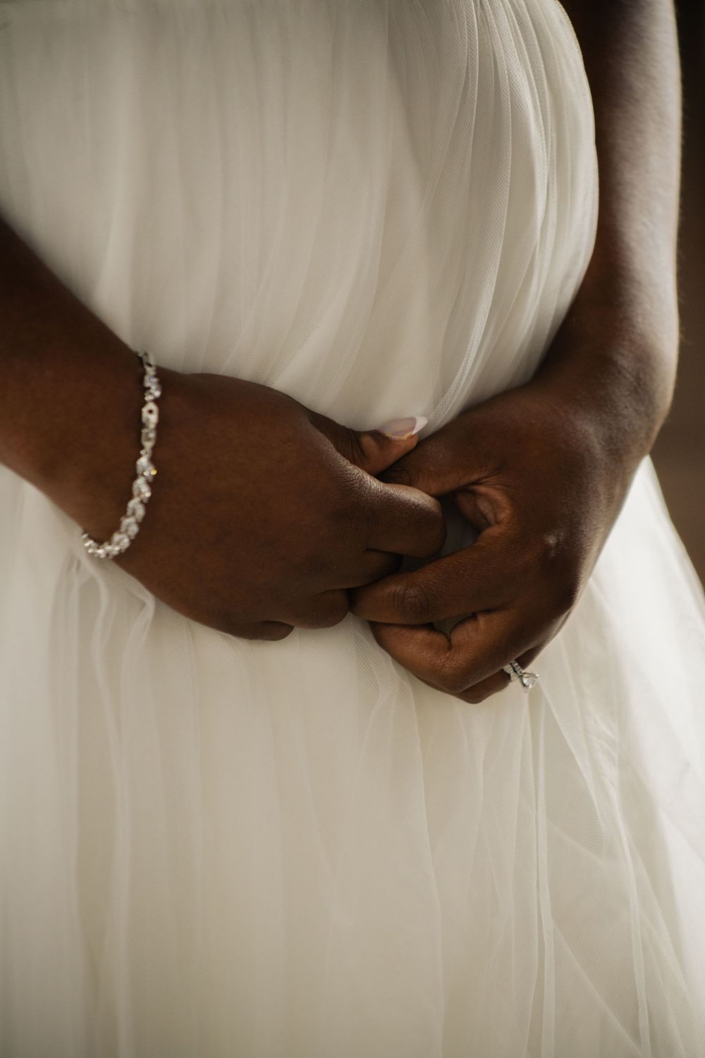 Close-up of bride's hands with delicate bracelet holding white wedding dress fabric.