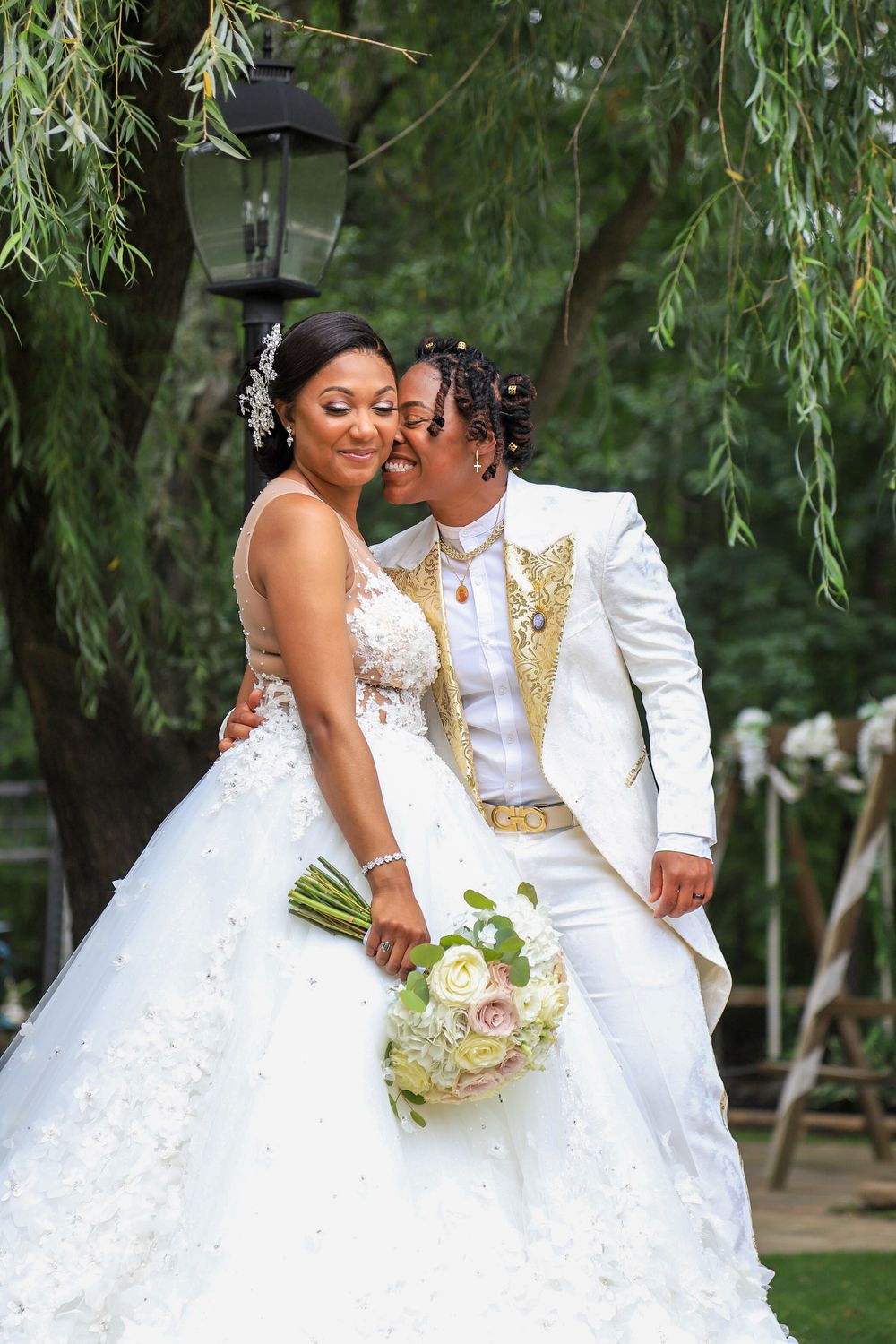 Wedding couple in white attire embrace under willow tree with romantic garden lighting.