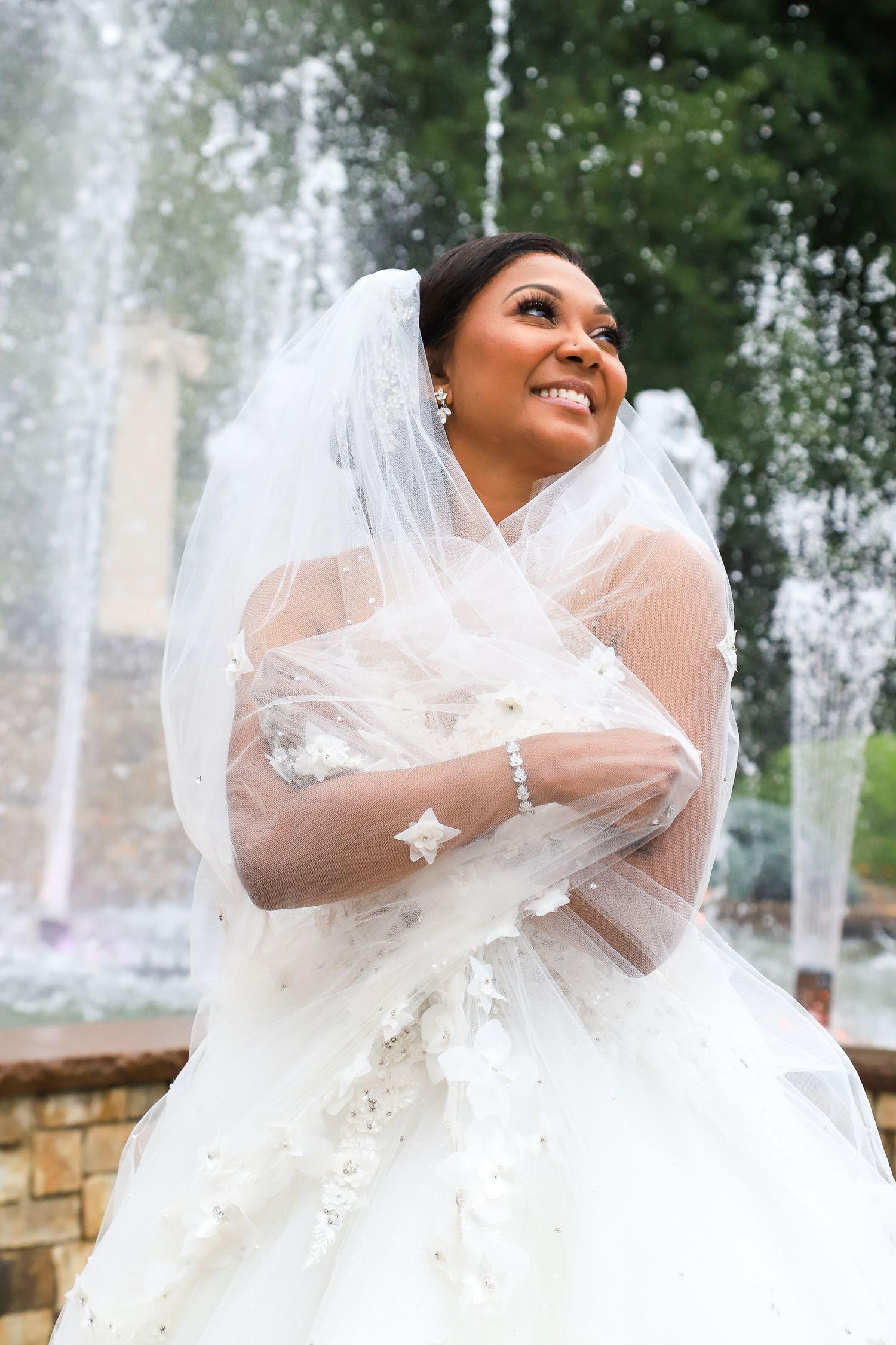 Radiant bride in lace gown and veil poses joyfully by fountain.