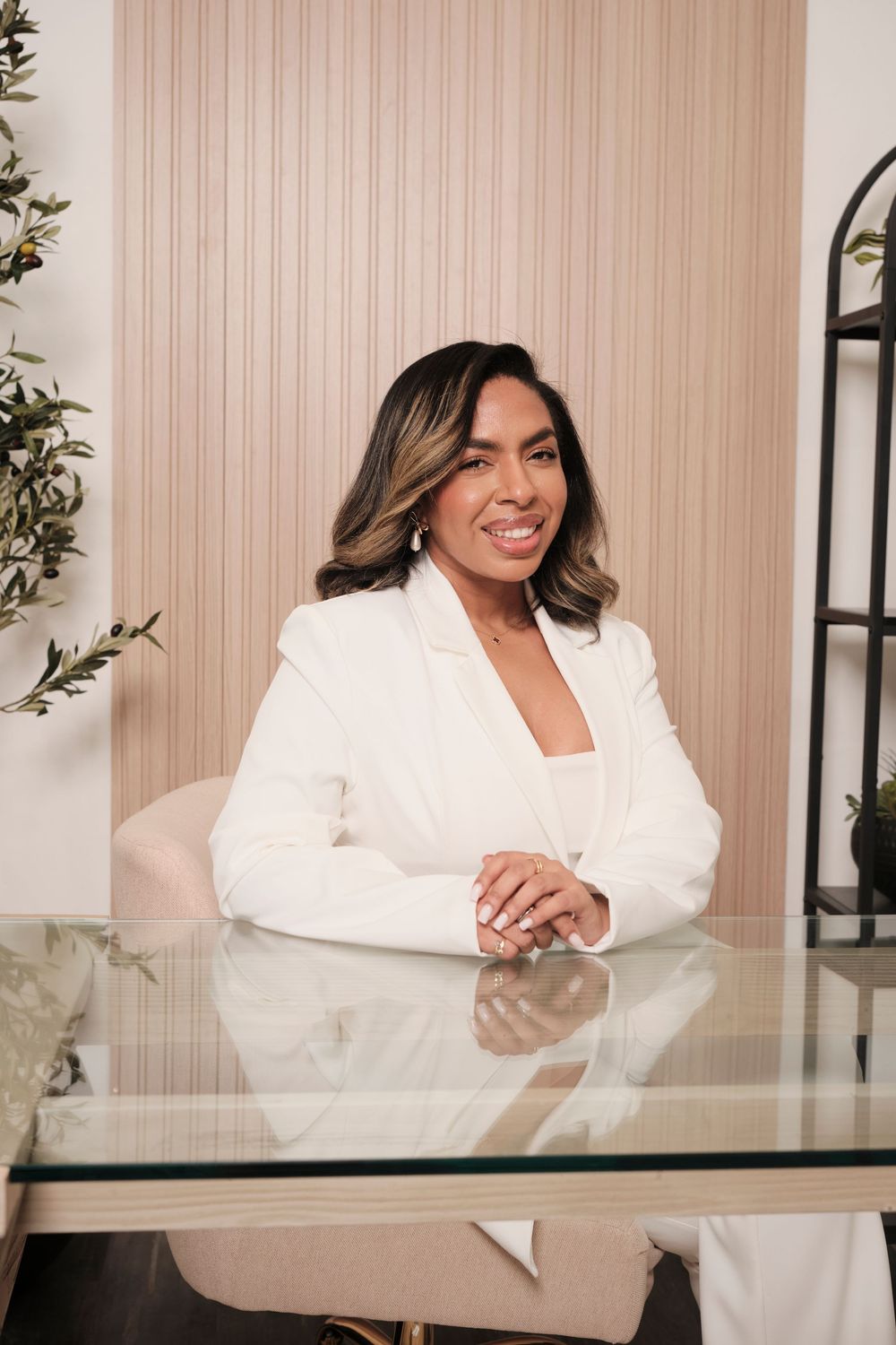 Professional in white blazer smiling while seated at glass desk in modern office setting.