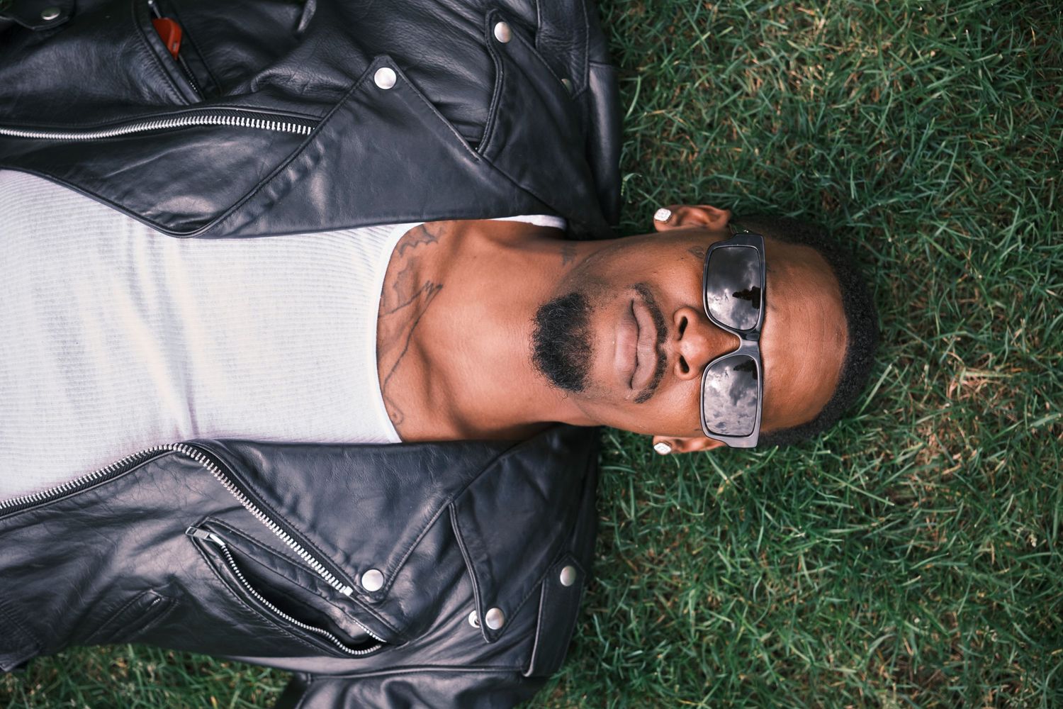 Man with sunglasses and beard lying on green grass wearing a leather jacket, relaxing outdoors.
