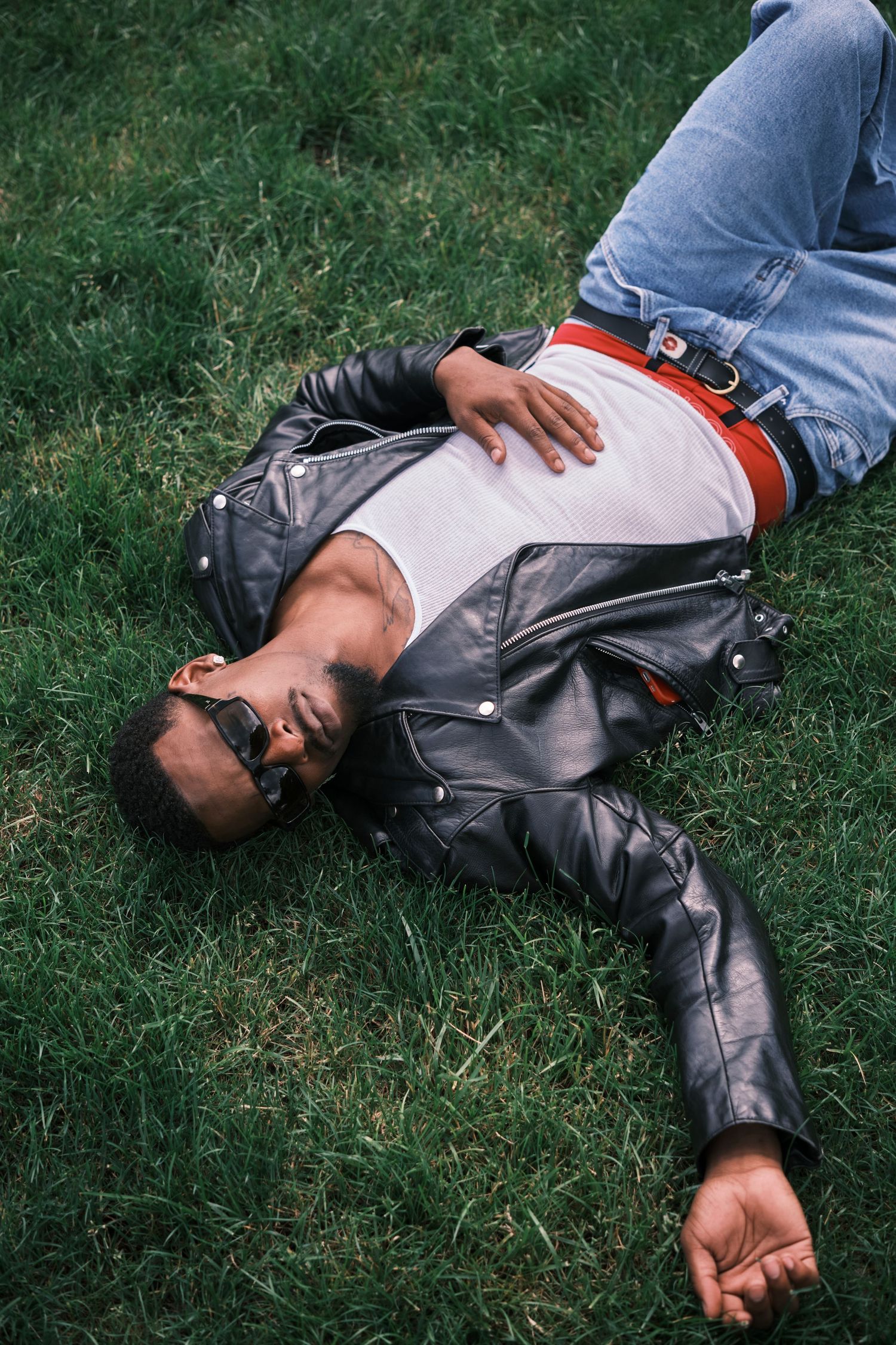 Man in leather jacket, jeans, and sunglasses relaxing on green grass outdoors.