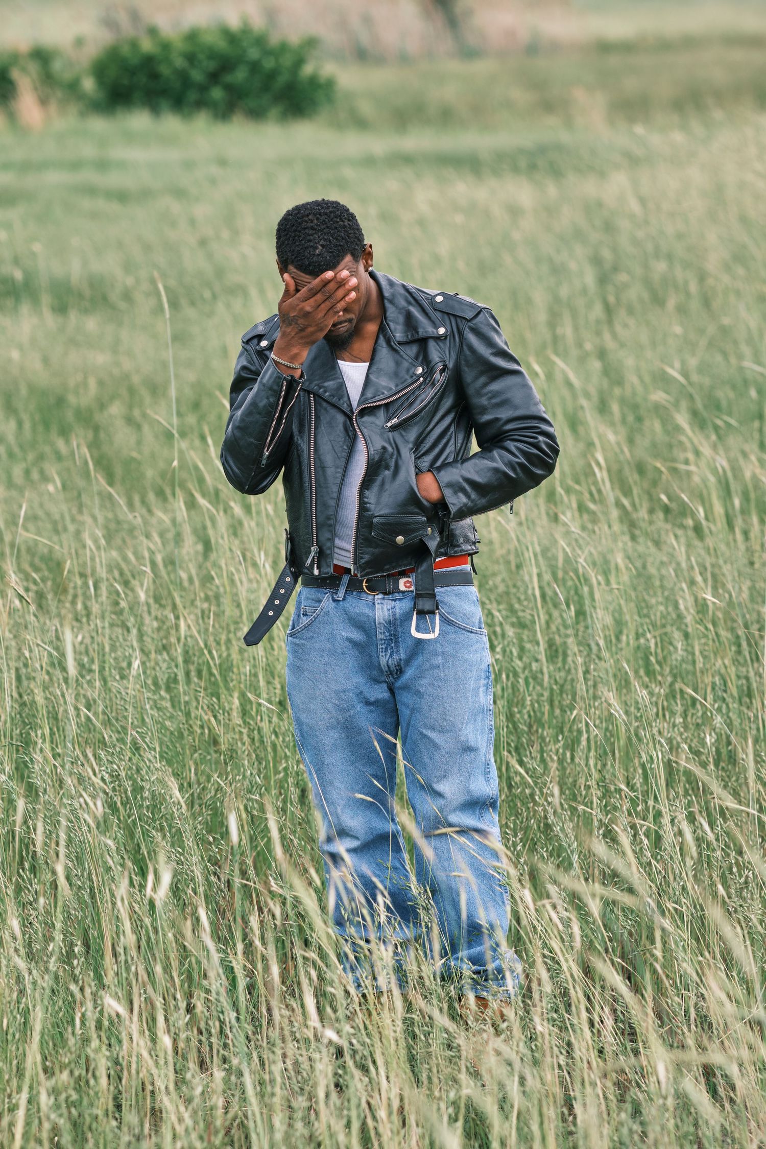 Black man in leather jacket and jeans standing in tall grass field, covering face with hand.