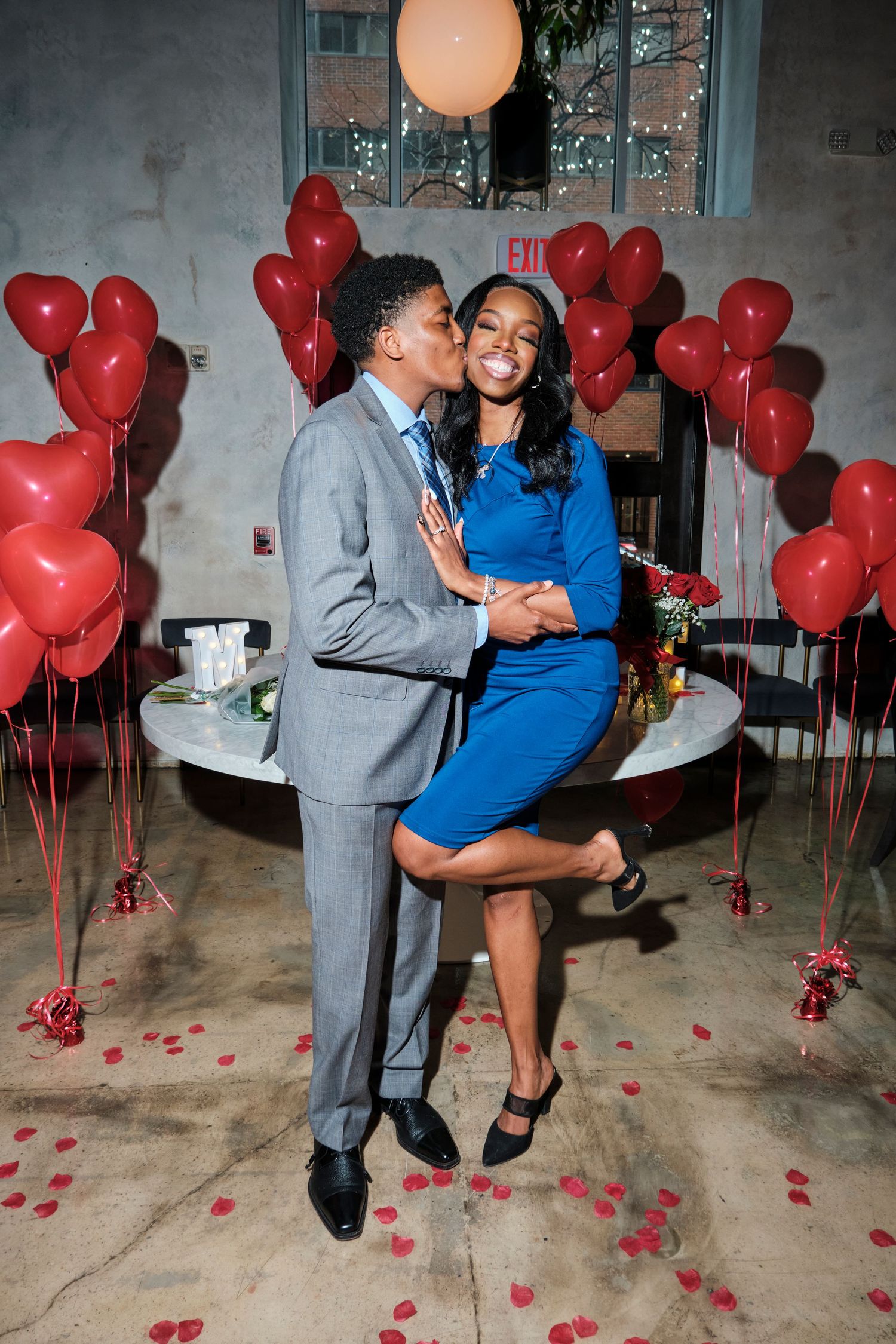 Couple dressed in blue dress and gray suit kissing in front of red heart balloons on Valentine's Day.