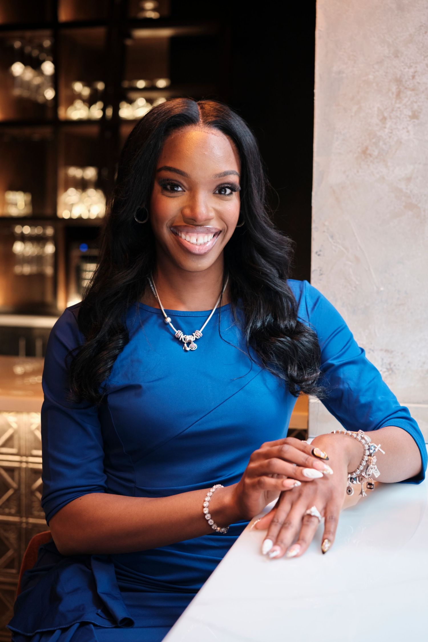 Smiling woman in blue dress seated at white table, wearing silver jewelry, in elegant restaurant setting.
