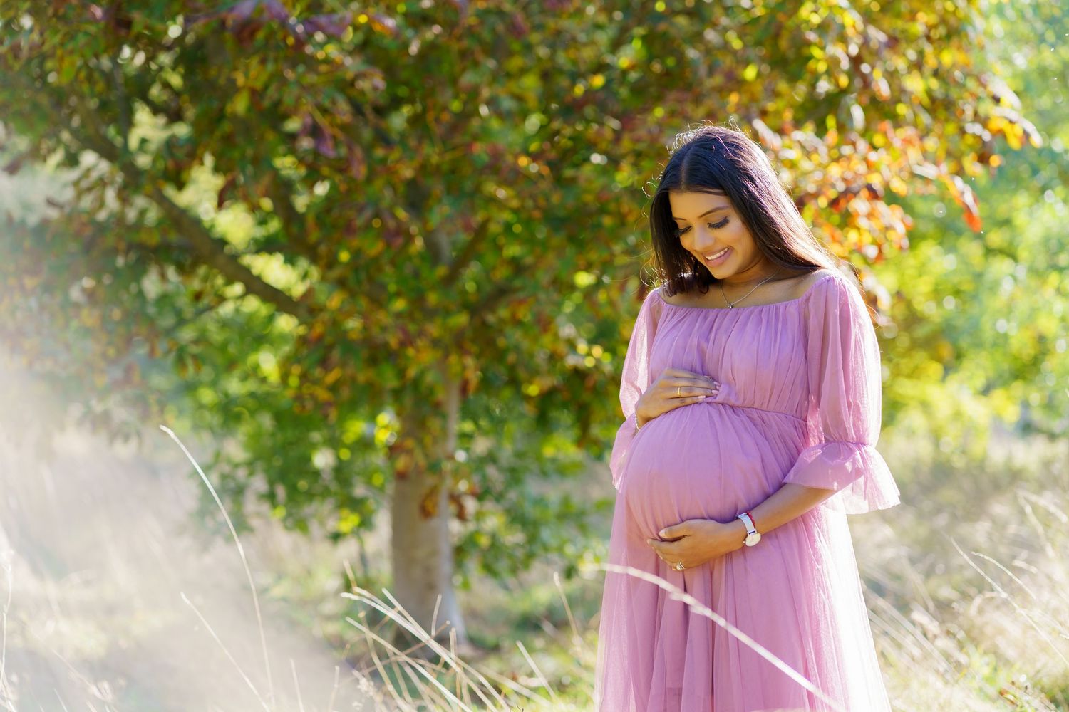 An expectant mother in a flowing pink dress stands in a sunlit garden with autumn foliage in the background.