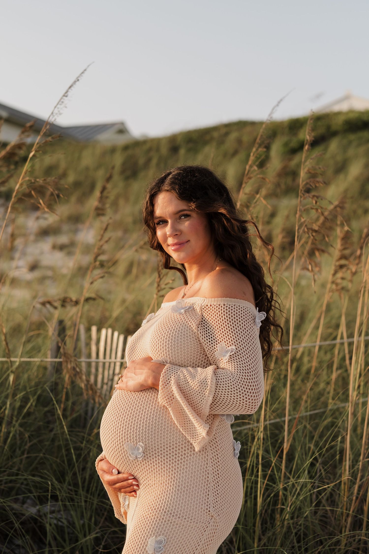Maternity photo of woman in cream dress posing among beach grass at sunset.
