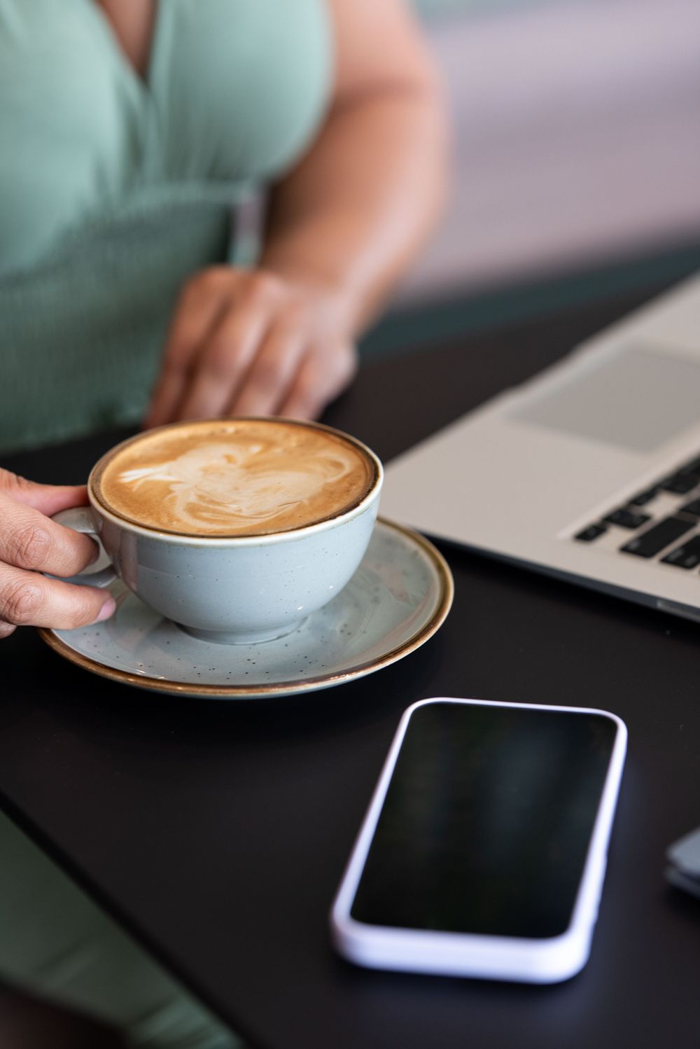 Latte art coffee cup on saucer next to smartphone and laptop on dark table.