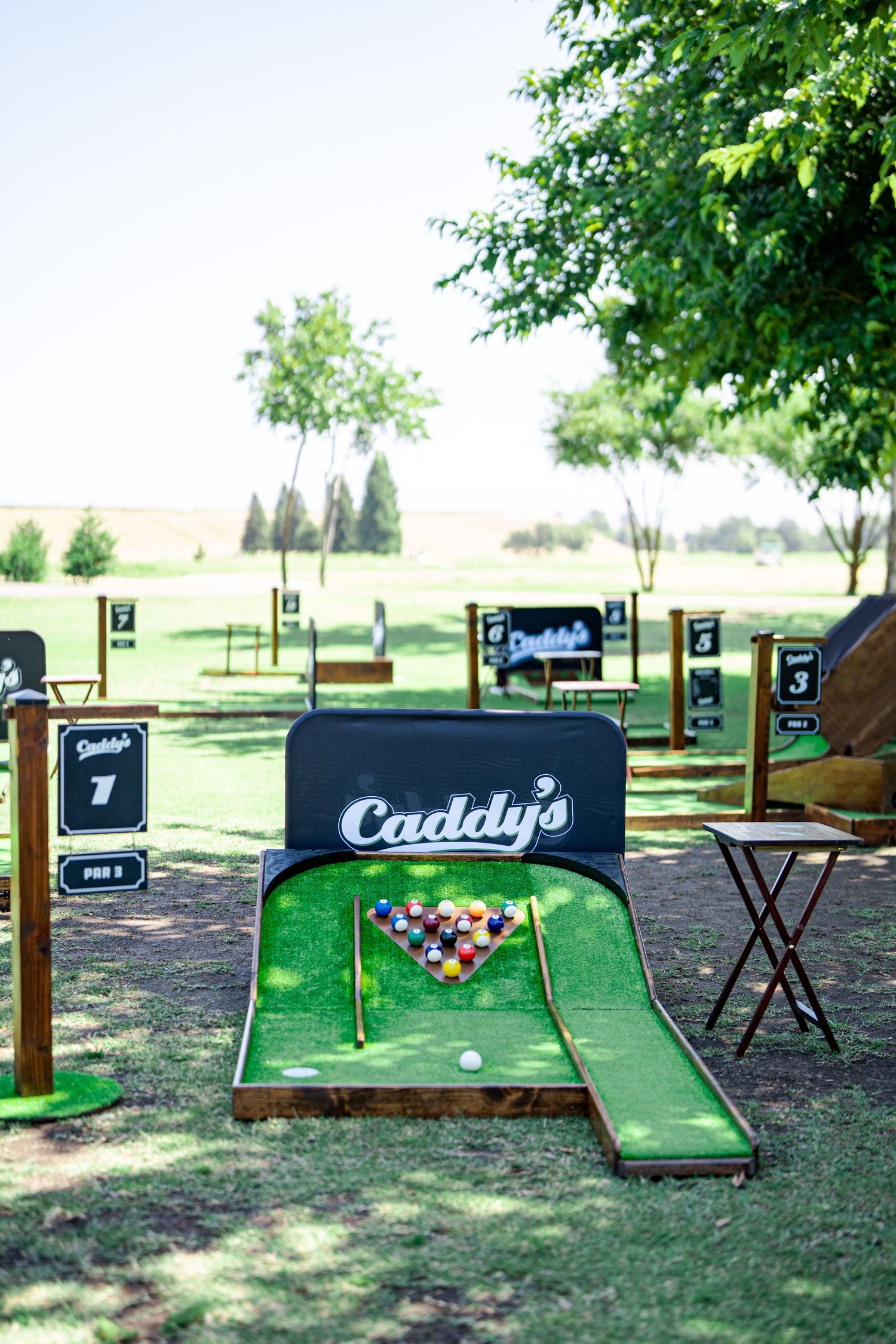 A series of miniature golf holes with wooden borders arranged on a grassy lawn in a backyard setting.