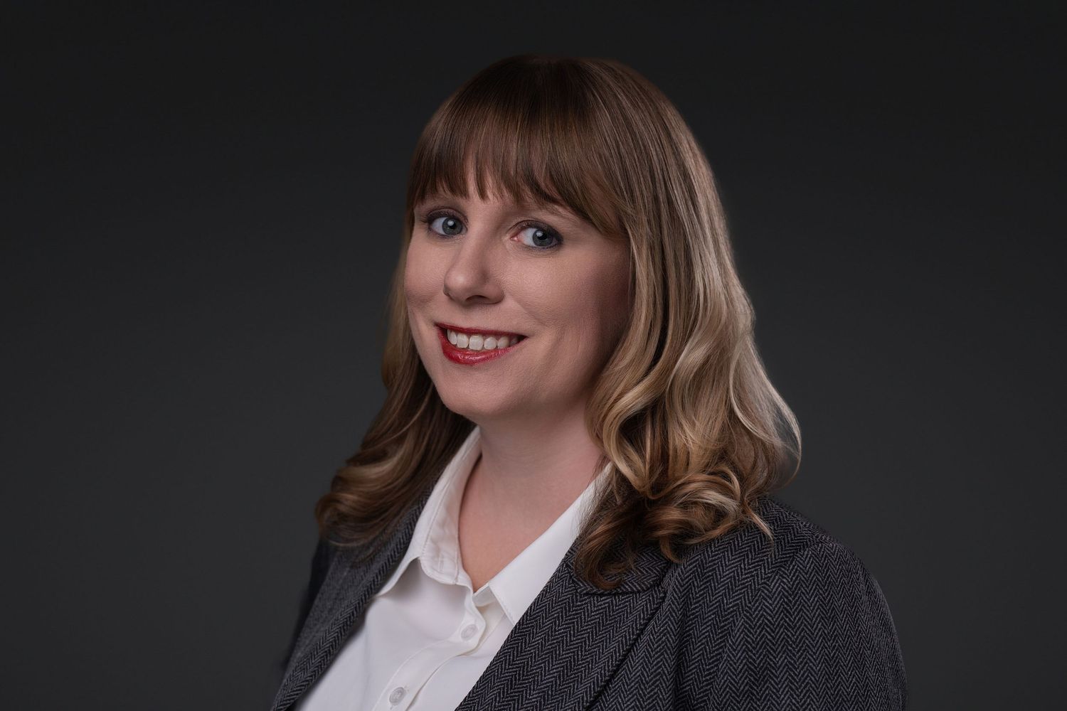 Professional woman with blonde bangs wearing a gray blazer and white blouse against a dark background.