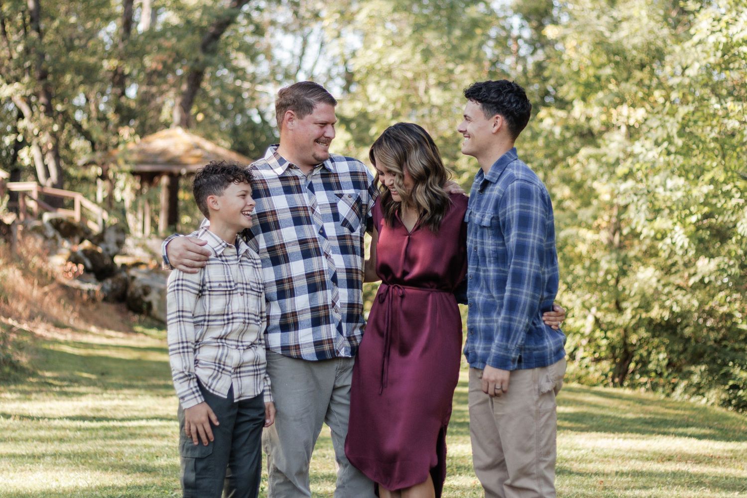 Four people in plaid and burgundy outfits smile together while standing in a sunny garden path.