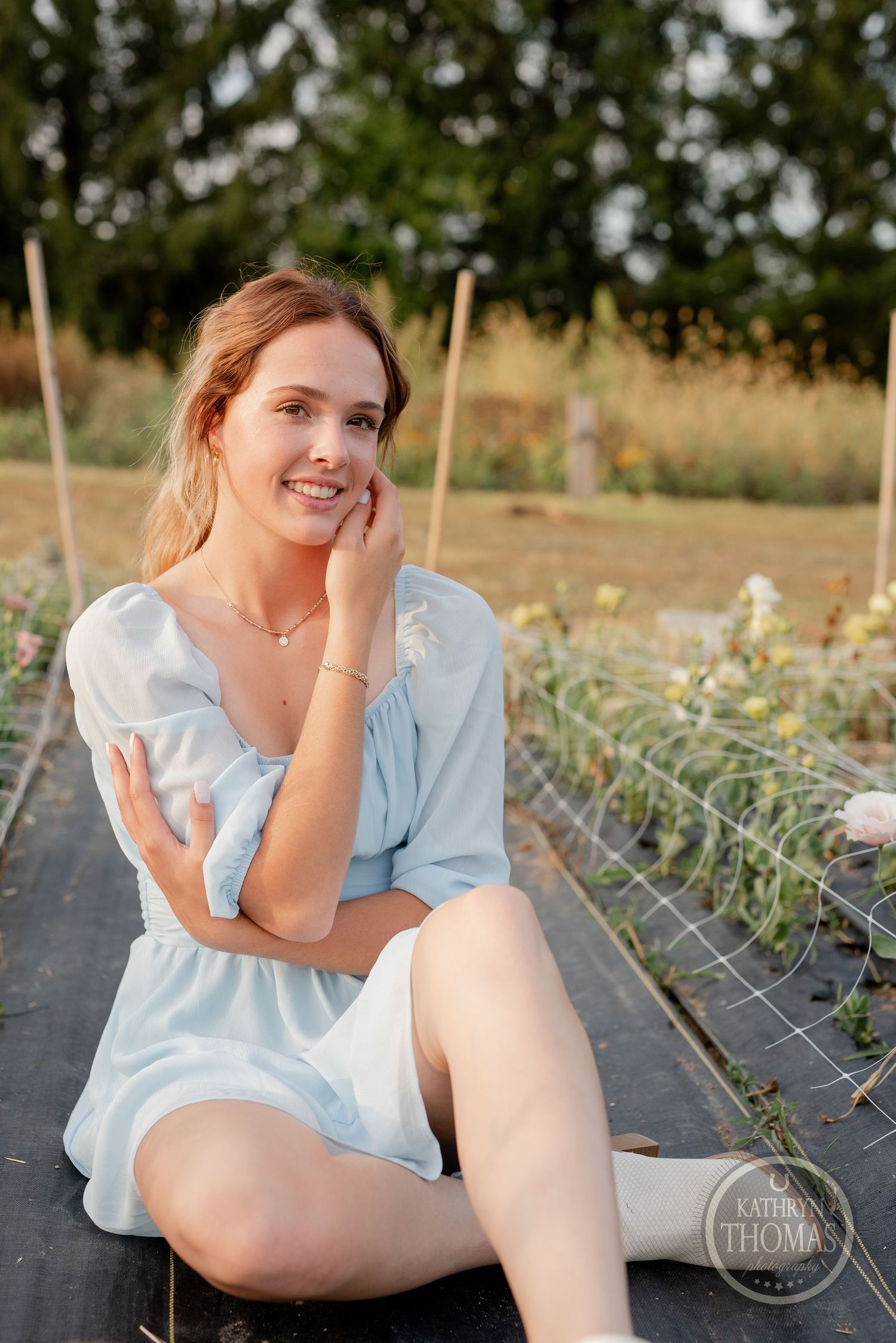 Someone in a light summer dress poses casually on a garden path with greenery in the background.