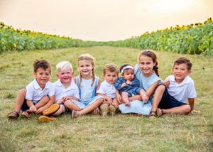 A cheerful group sits together on grassy ground between rows of tall sunflowers.