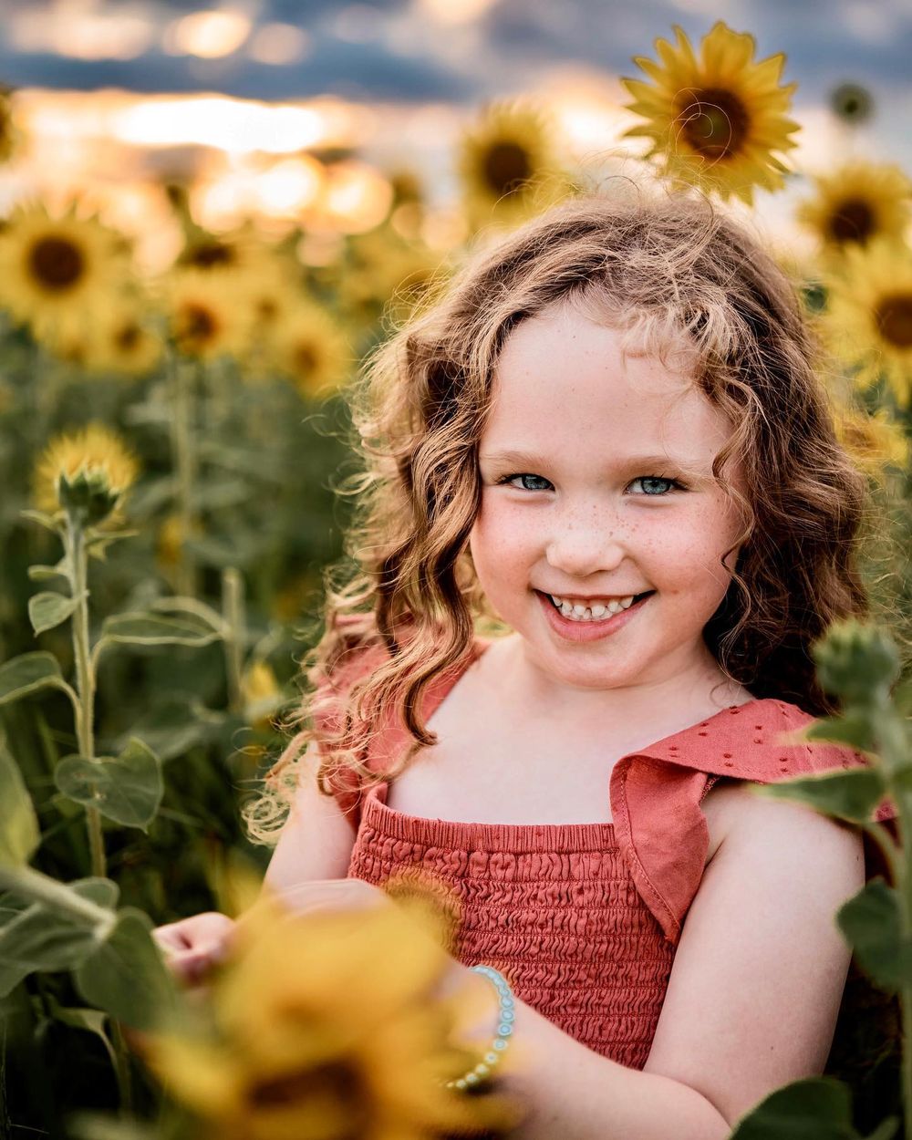 Young girl smiling in sunflower field wearing coral dress.