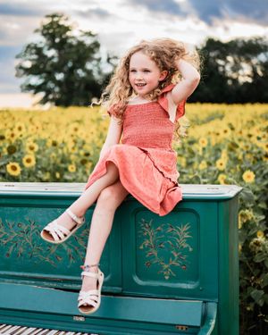 Girl in coral dress sitting playfully on turquoise piano amid sunflowers.