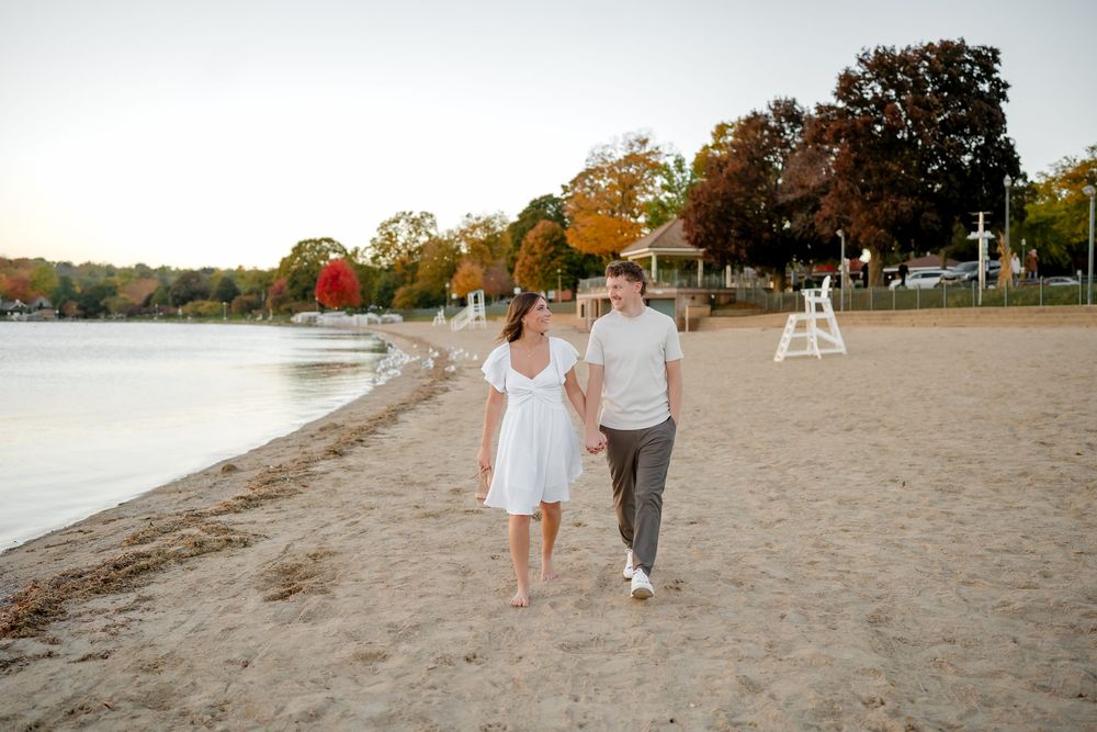 A couple walks hand in hand along a sandy beach at sunset with fall foliage and a lifeguard stand in the background.