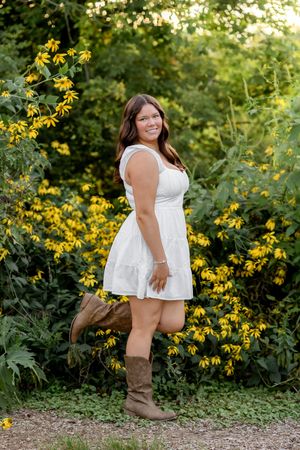Someone in a white sundress and brown boots poses among blooming yellow wildflowers on a summer evening.