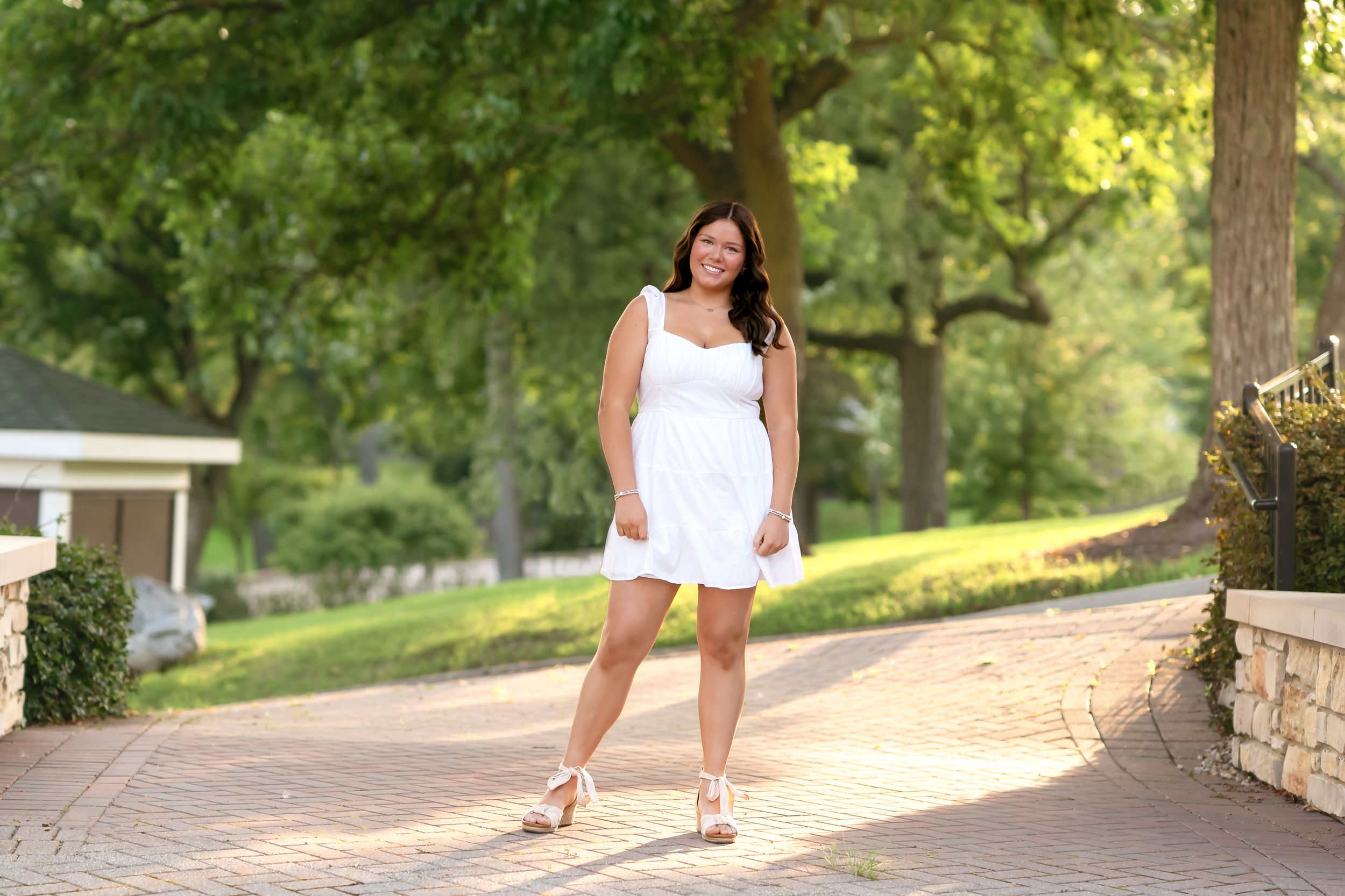Woman in white dress walking along sunlit stone pathway through lush green garden setting.