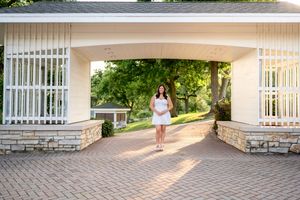 Person in a white dress stands beneath an elegant stone archway with white columns on a brick path at sunset.