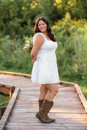 Person in a white summer dress and brown boots poses playfully on a wooden boardwalk surrounded by greenery.