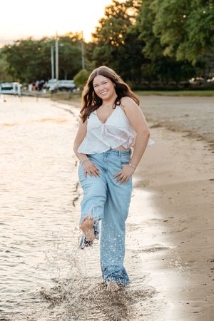 Person in denim and white top splashes joyfully in shallow water at the beach during golden hour.